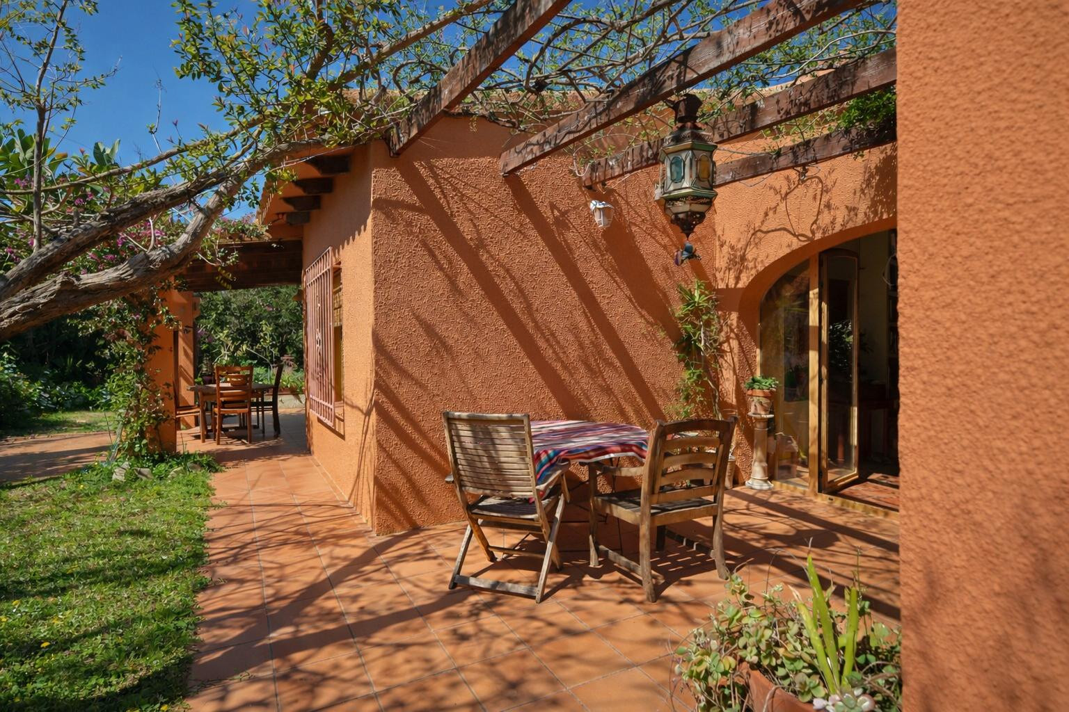 Buitenterras met terracotta tegels, een houten pergola met klimplanten en rustiek houten meubilair tegen een warmgekleurde gevel.