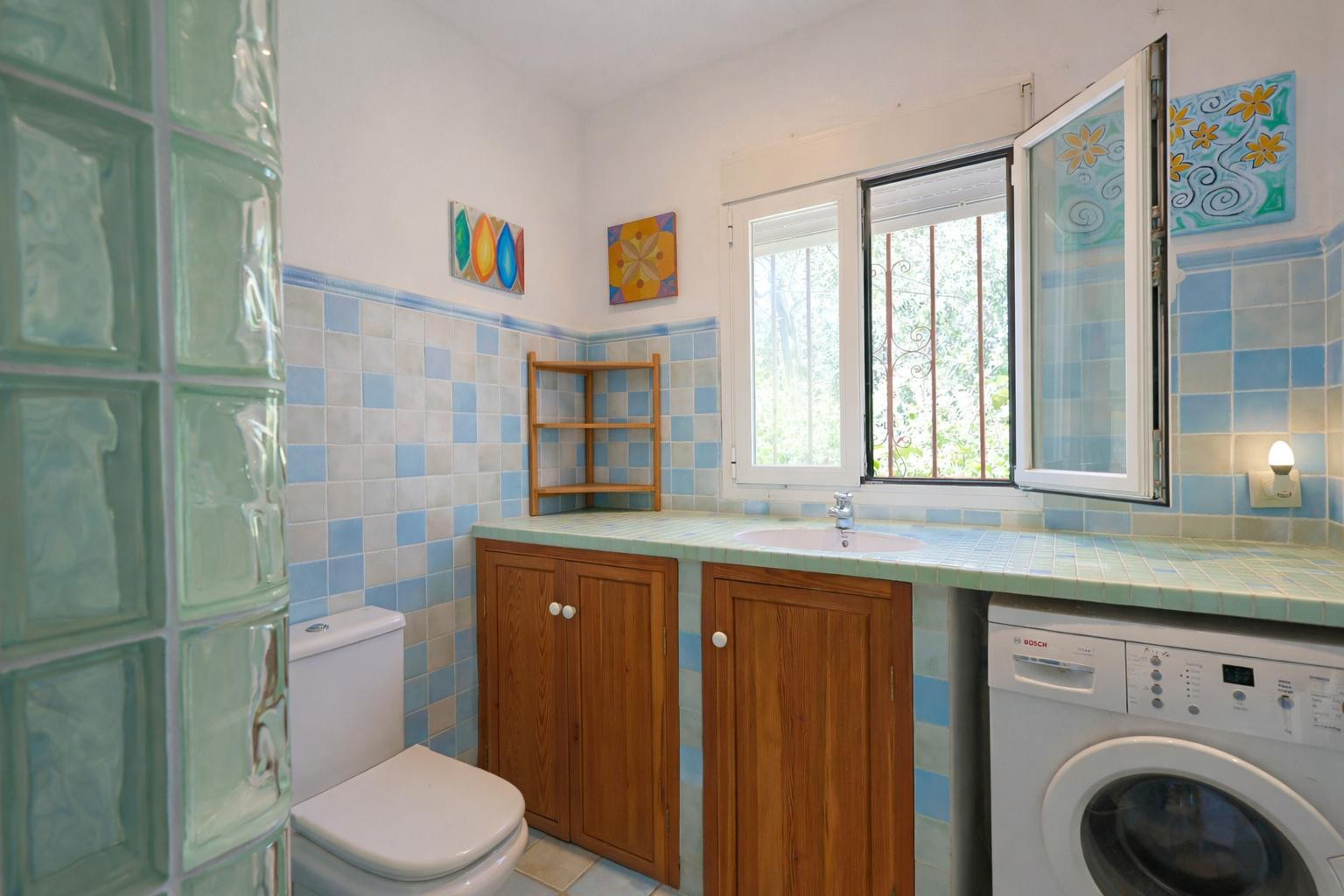 Functionele badkamer met blauwe betegeling, een betegeld werkblad met geïntegreerde wastafel, houten kasten en een wasruimte met daglicht.