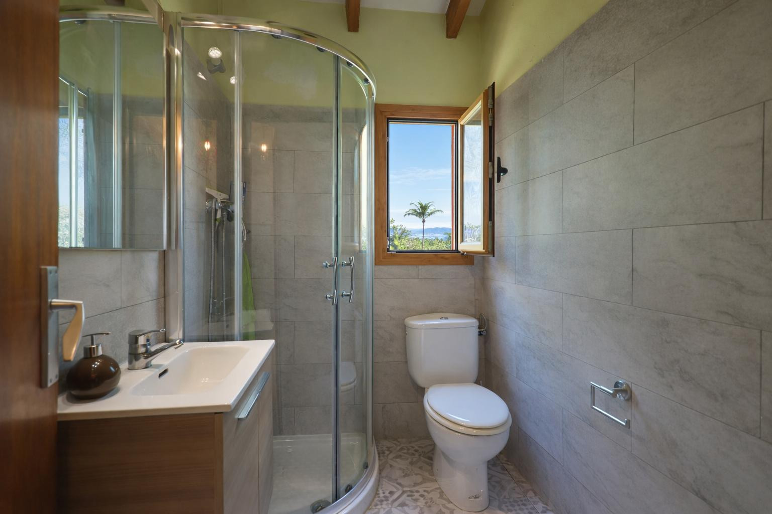 Functionele badkamer met een ronde glazen douchecabine, grijze tegels en houten plafondbalken. Het raam zorgt voor natuurlijk licht en uitzicht op de omgeving.