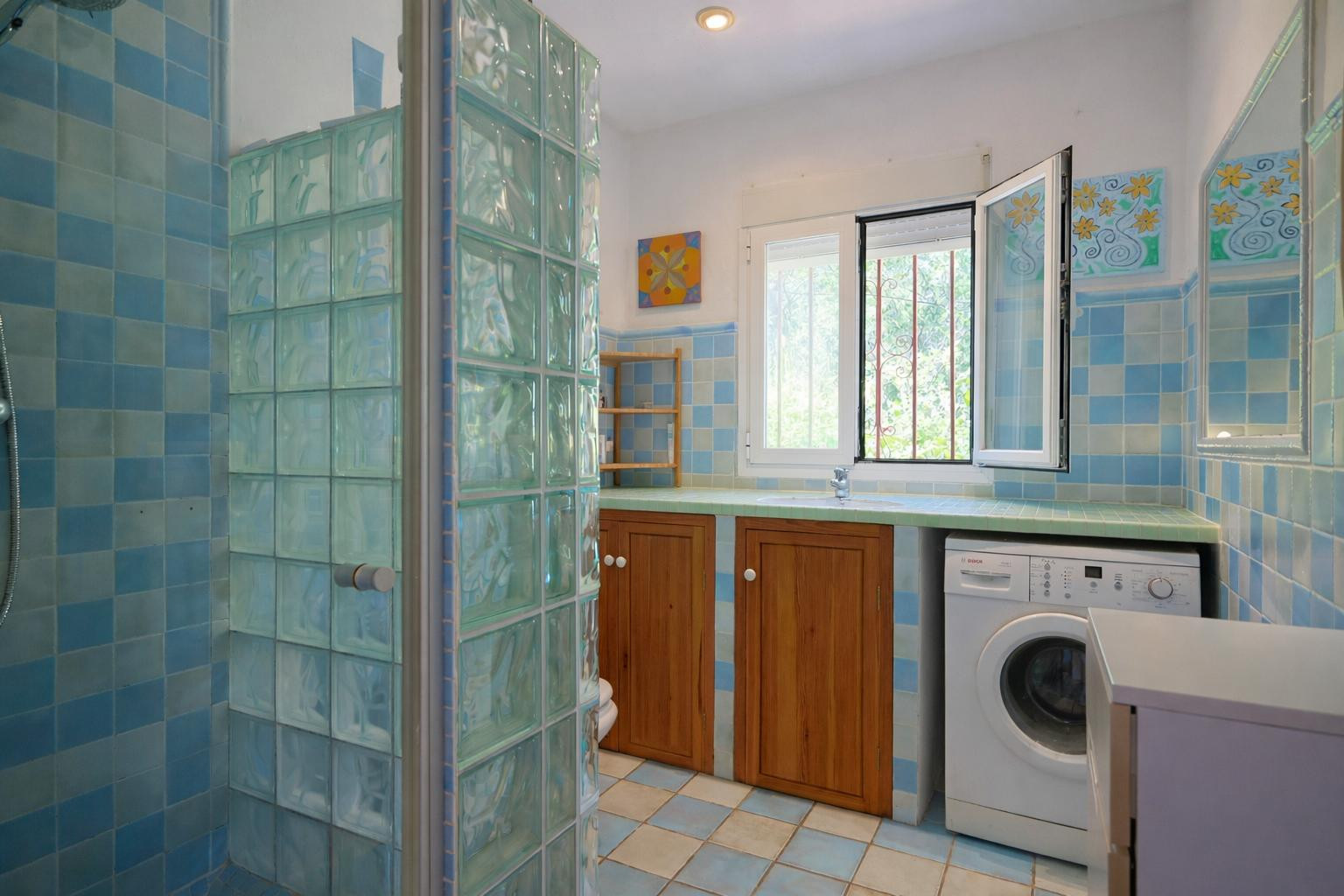 Functionele badkamer met wasruimte, blauwe dambordtegels, glazen bouwstenen douchewand, houten kasten en een geïntegreerde wasmachine.