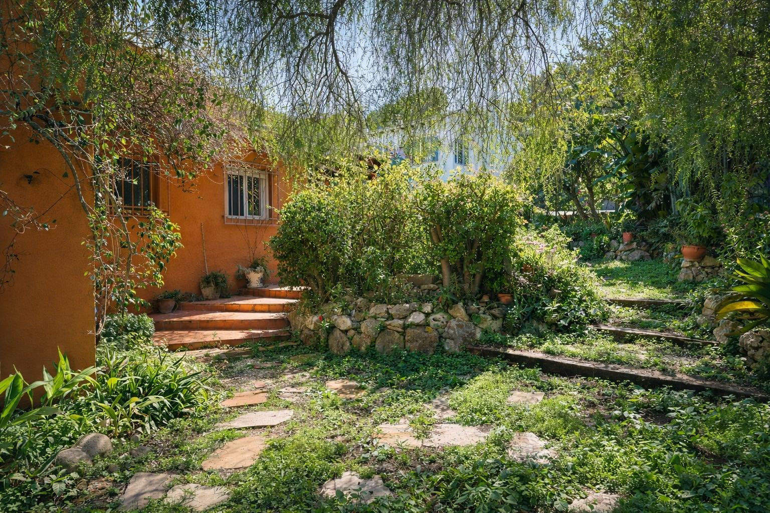 Mediterrane tuin met stenen paden en houten treden, omgeven door volwassen begroeiing en natuurstenen muren tegen de terracotta gevel.