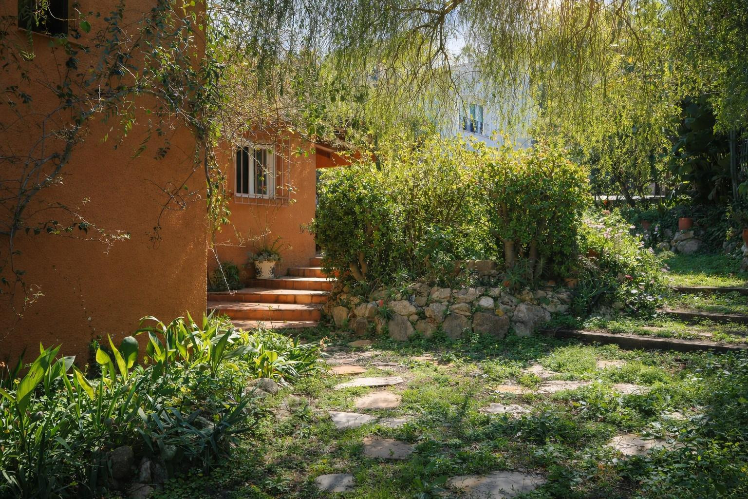 Natuurlijke tuin met stenen paden en terracotta treden die naar de okerkleurige gevel van de villa leiden, omgeven door volgroeide mediterrane beplanting.