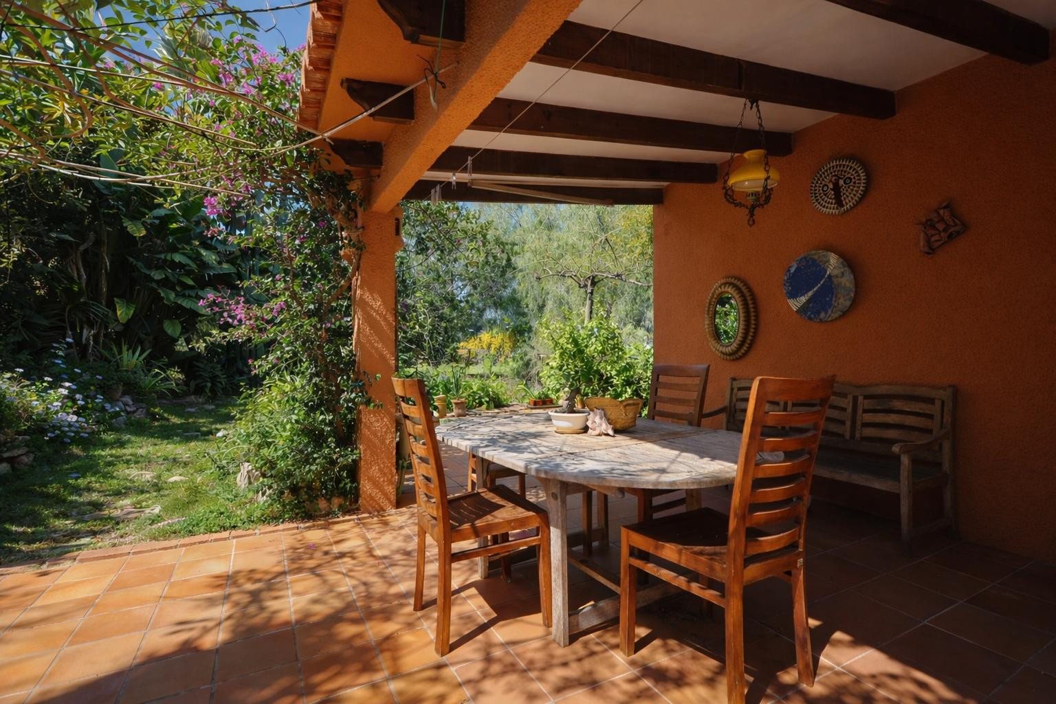 Overdekt terras met houten balkenplafond, terracotta tegels en een eethoek buiten met uitzicht op de volgroeide mediterrane tuin.