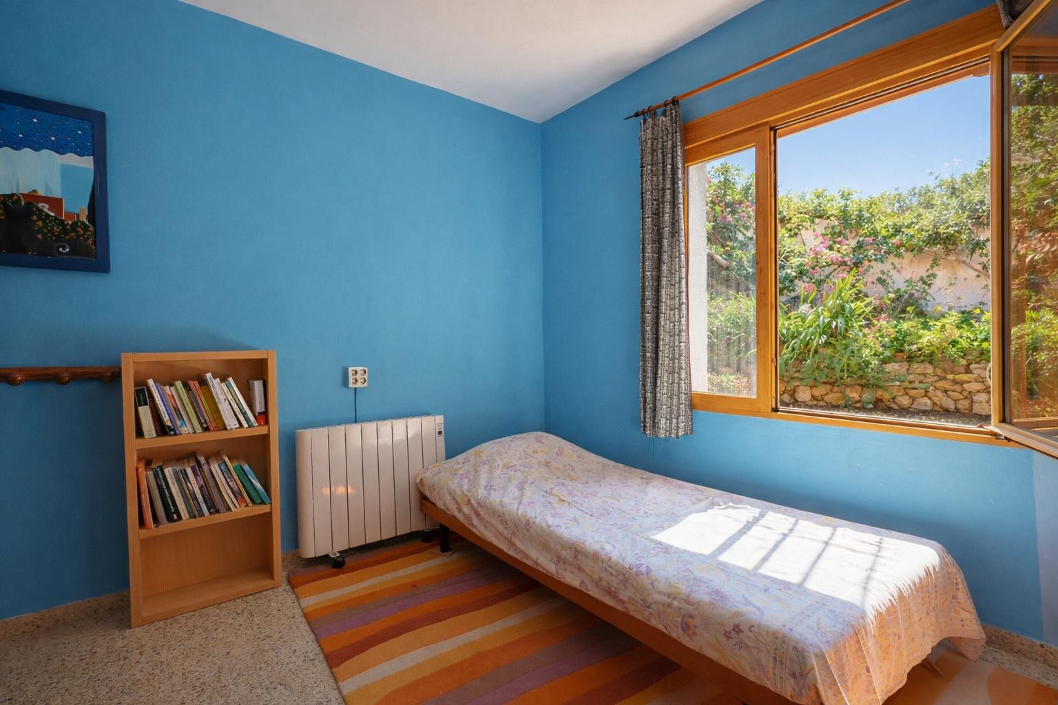 Slaapkamer met blauwe wanden, een eenpersoonsbed en houten boekenkast. Het houten raamkozijn kijkt uit op de tuin met natuurstenen muren en beplanting.
