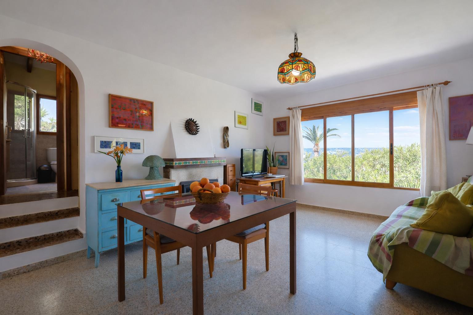 Woon- en eetkamer met open haard, terrazzovloer en een groot houten raam met uitzicht op zee. Een boog biedt toegang tot de nabijgelegen badkamer.