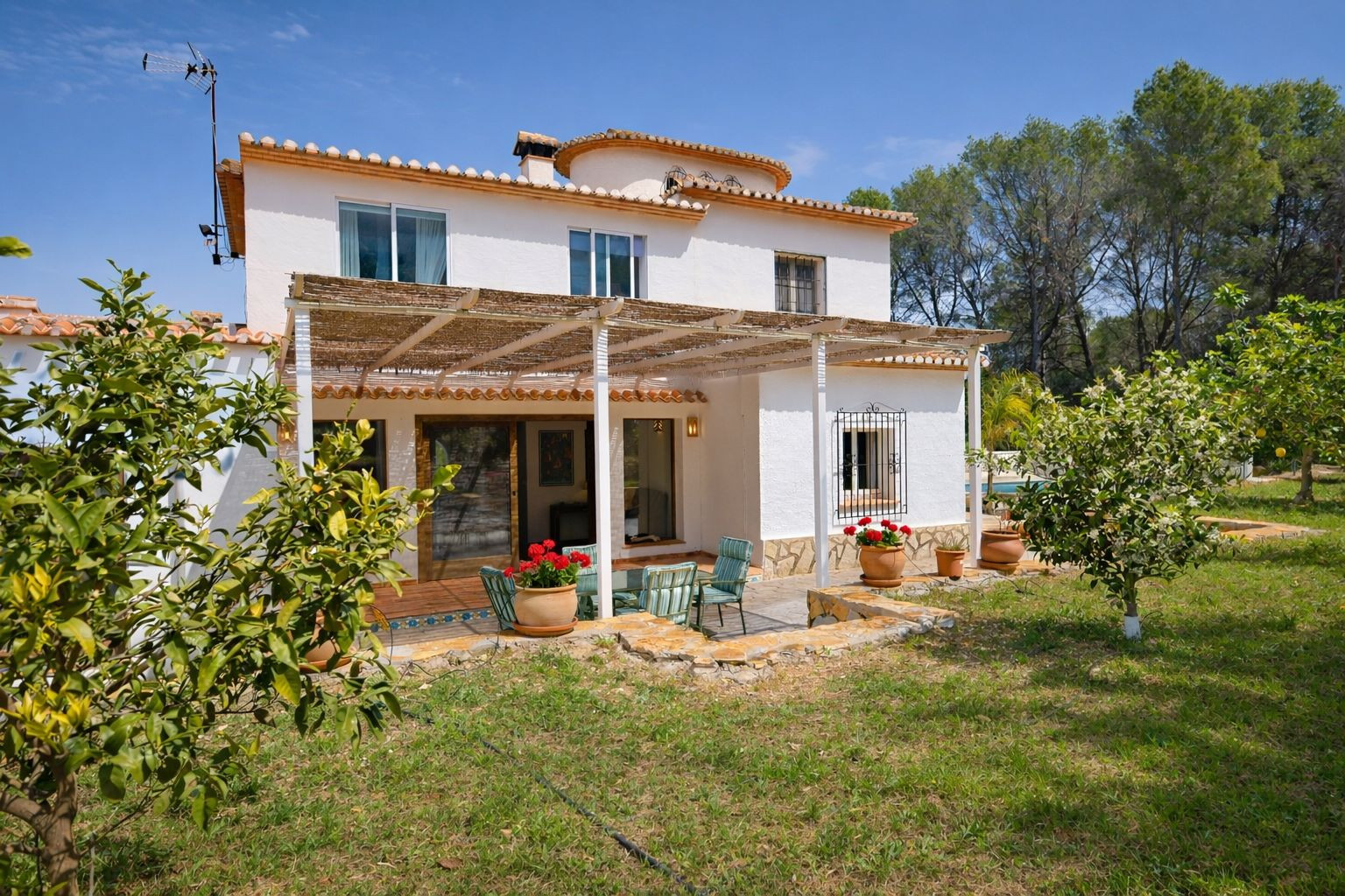 Achtergevel van de villa met een rietgedekt terras, zithoek buiten en een tuin met fruitbomen. Mediterrane architectuur met stenen details en terracotta dakpannen.