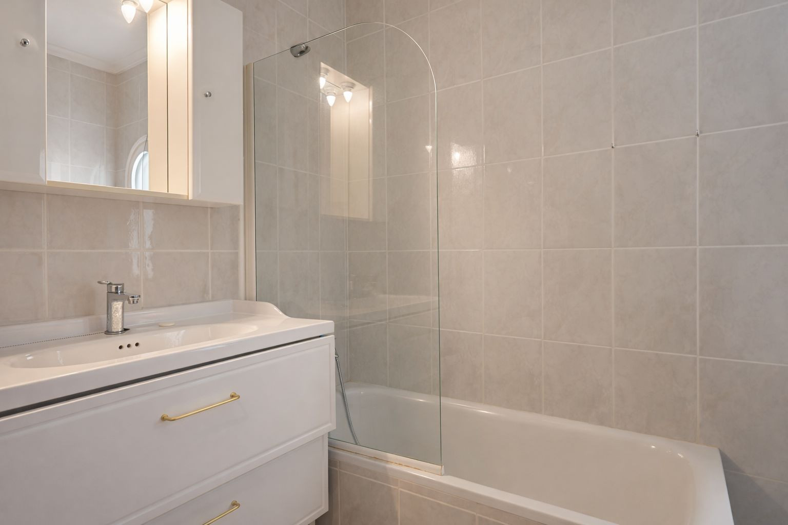 Badkamer voorzien van een ligbad met gebogen glazen wand, wit wastafelmeubel met goudkleurige grepen en een spiegelkast met verlichting.