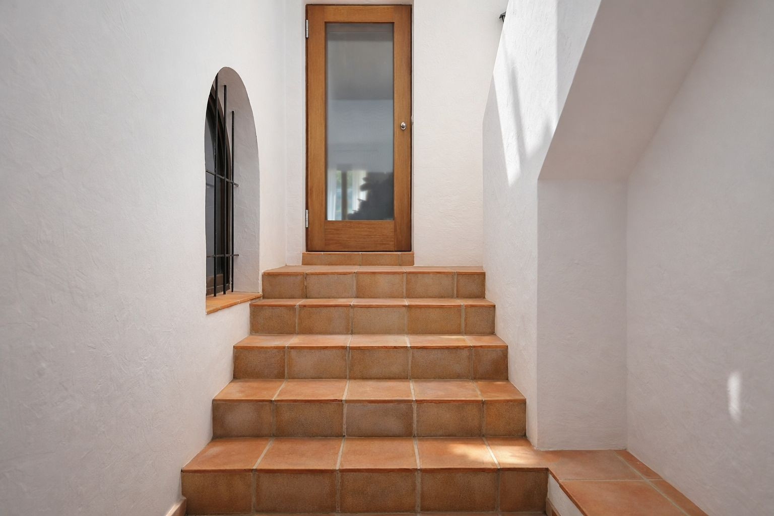 Binnentrap met terracotta treden die leidt naar een houten deur met glas, voorzien van een boogvenster met smeedijzeren tralies en witte wanden.