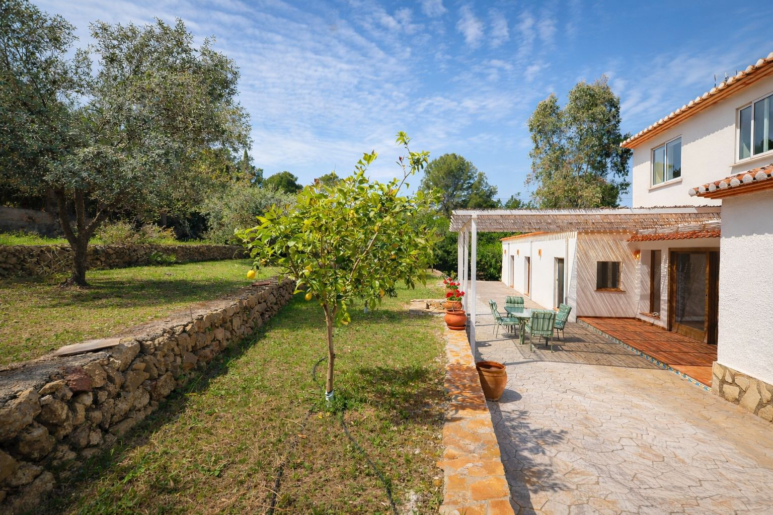 Buitenaanzicht met geplaveid terras, schaduwrijke pergola en terrasvormige tuin met natuurstenen muren en fruitbomen.