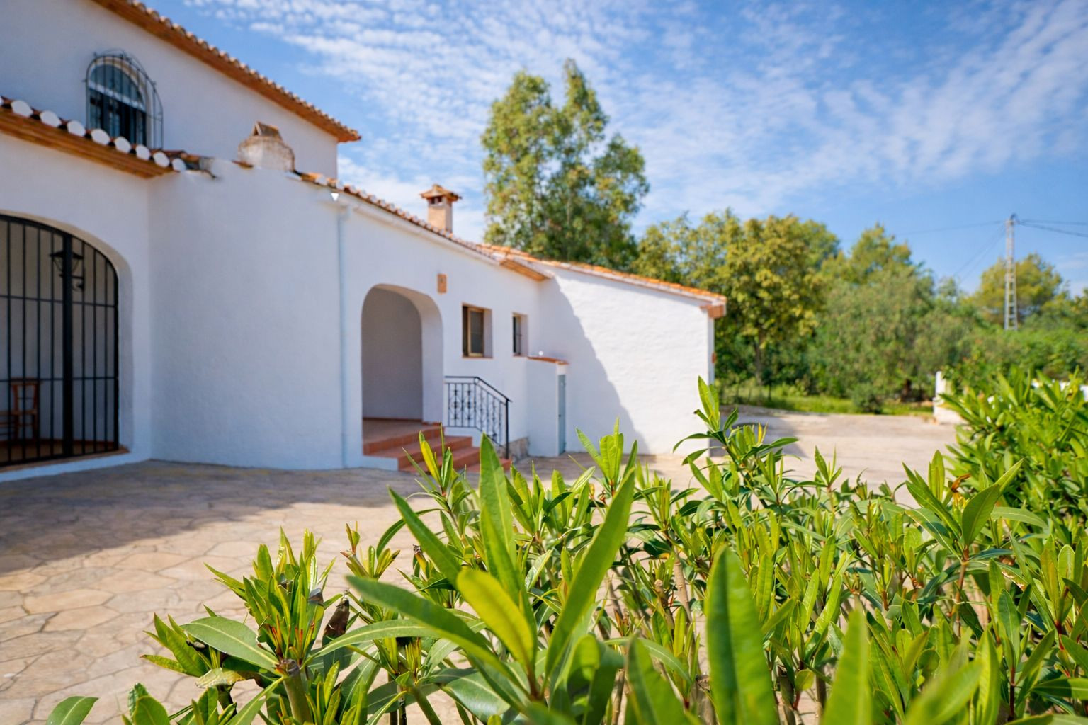 Buitenaanzicht van de witte villa met keramische dakpannen, een natuurstenen terras en boogvormige details, omgeven door mediterrane beplanting.