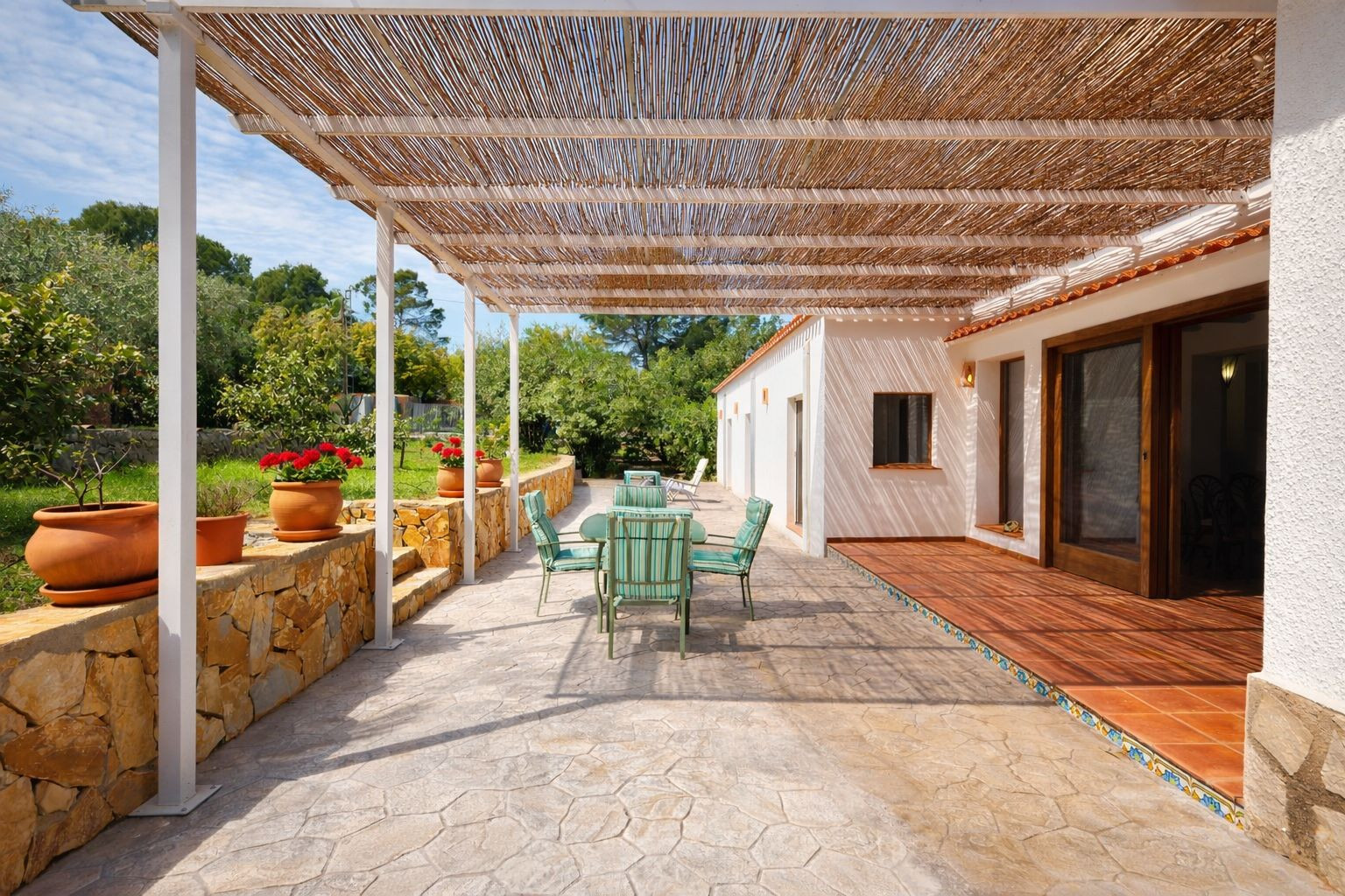 Buitenterras met een pergola van riet, natuurstenen vloer en tuinmeubilair. Een natuurstenen muur met terracotta potten grenst aan de omliggende tuin.