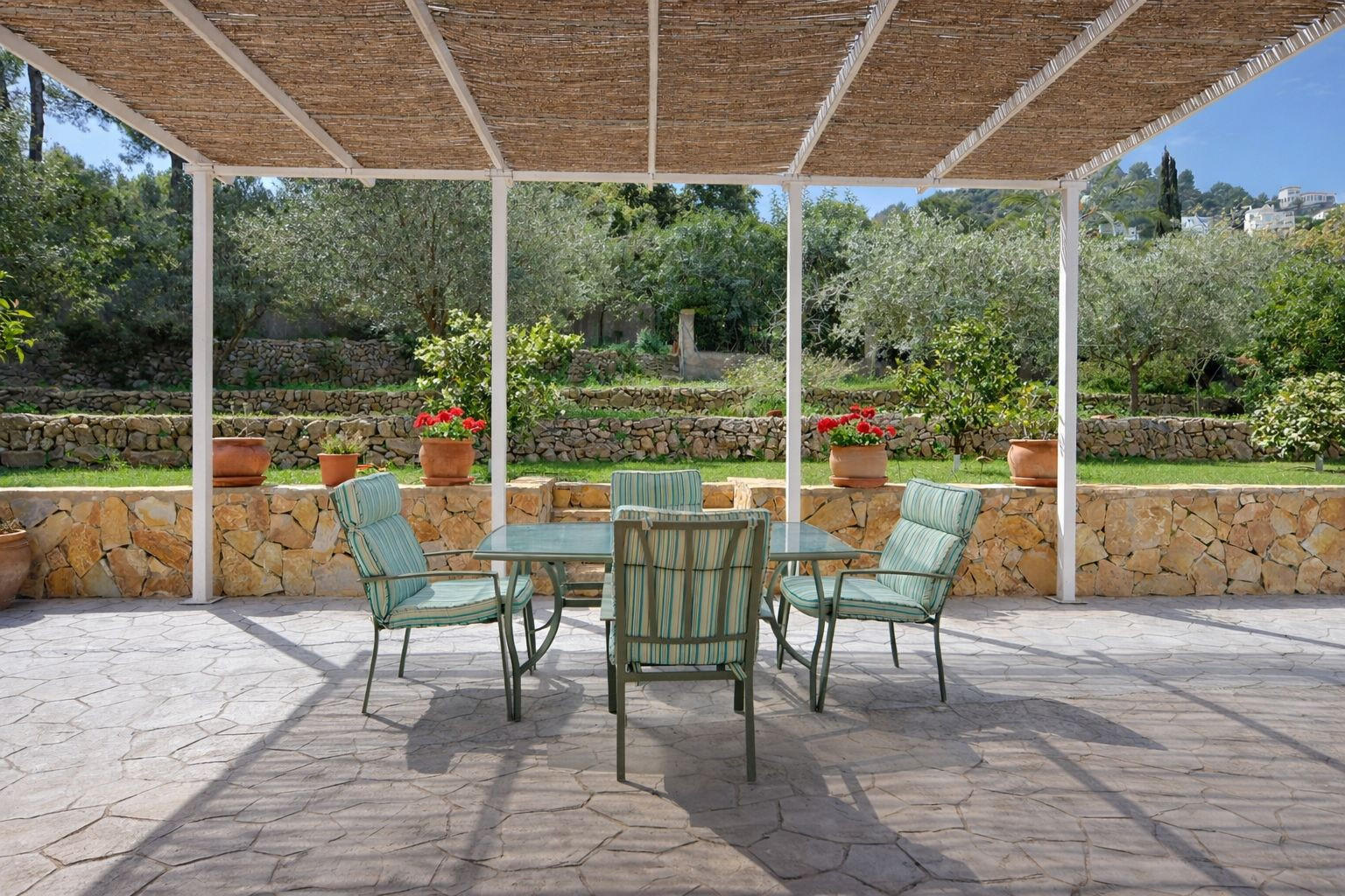 Buitenterras met een rieten pergola, eethoek en uitzicht op een terrasvormige tuin met natuurstenen muren en fruitbomen.