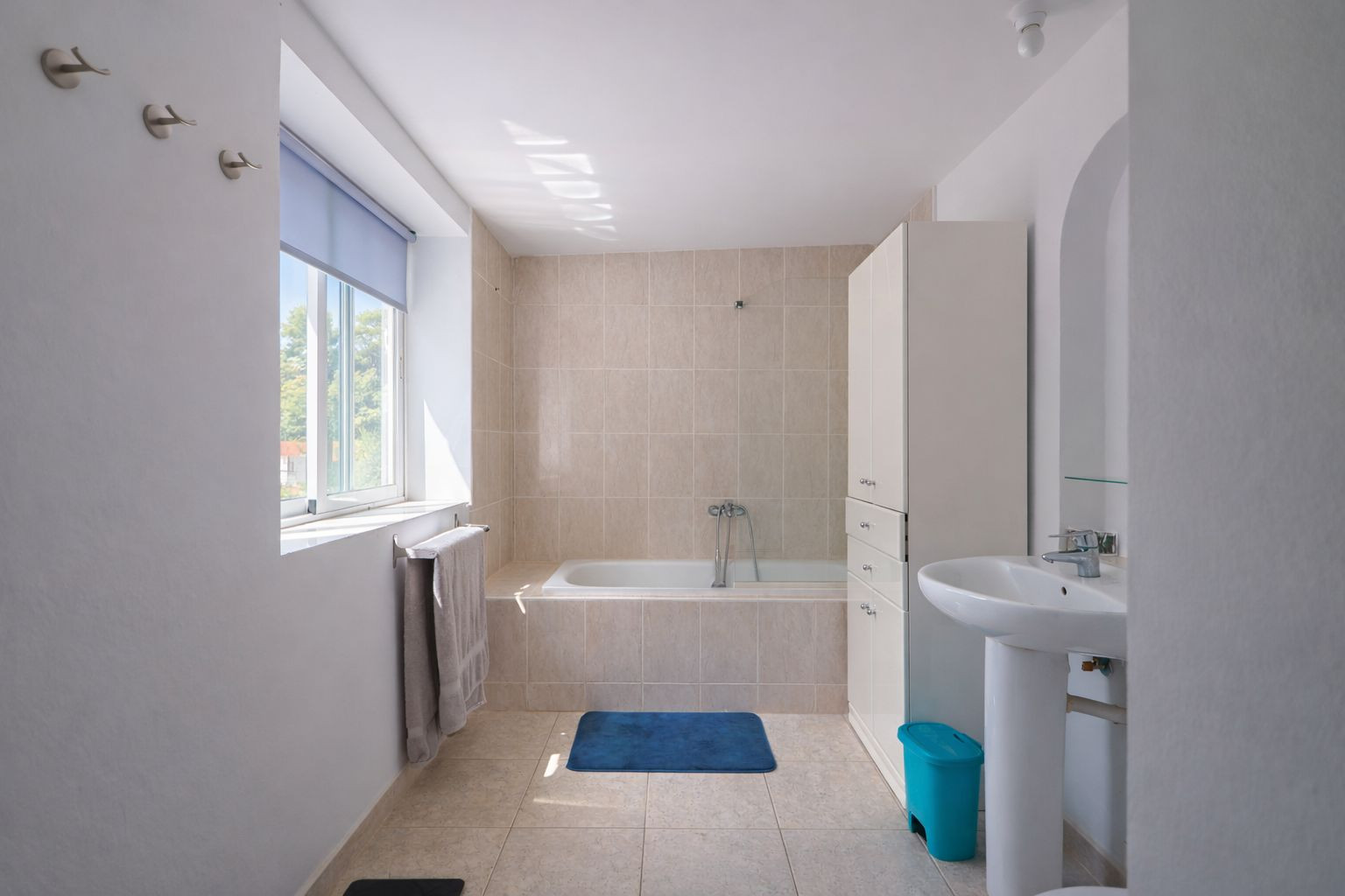 Functionele badkamer met ingebouwd ligbad, neutrale keramische tegels, een kolomwastafel en een raam voor natuurlijk licht.