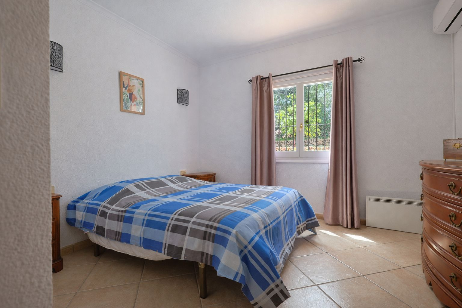 Lichte slaapkamer met tegelvloer, klassiek houten meubilair en uitzicht op de tuin. Voorzien van airconditioning en elektrische wandverwarming.