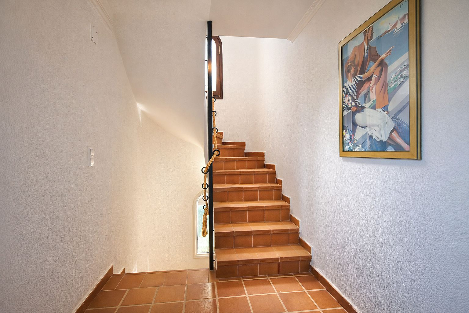 Trap met terracotta tegels en een zwart metalen leuning met touwdetail, omgeven door witte muren en natuurlijk licht via een klein raam.