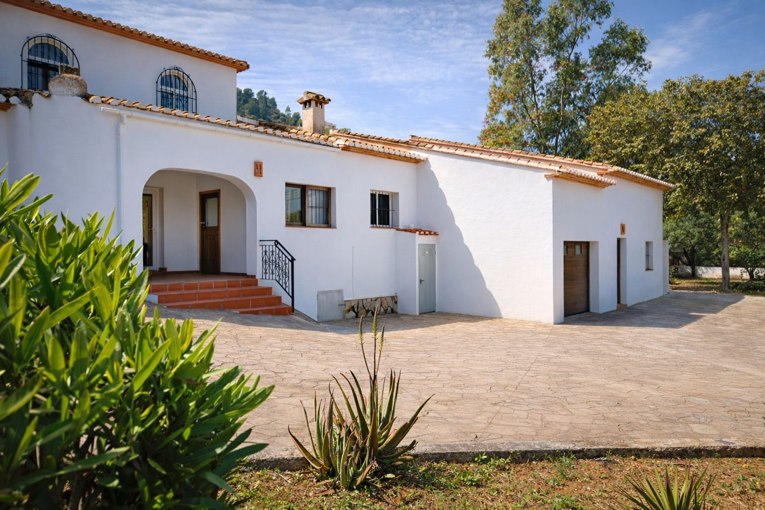 Witte gevel in mediterrane stijl met terracotta dakpannen, een gewelfde entree en een ruim geplaveid voorterrein dat toegang biedt tot de inpandige garage.