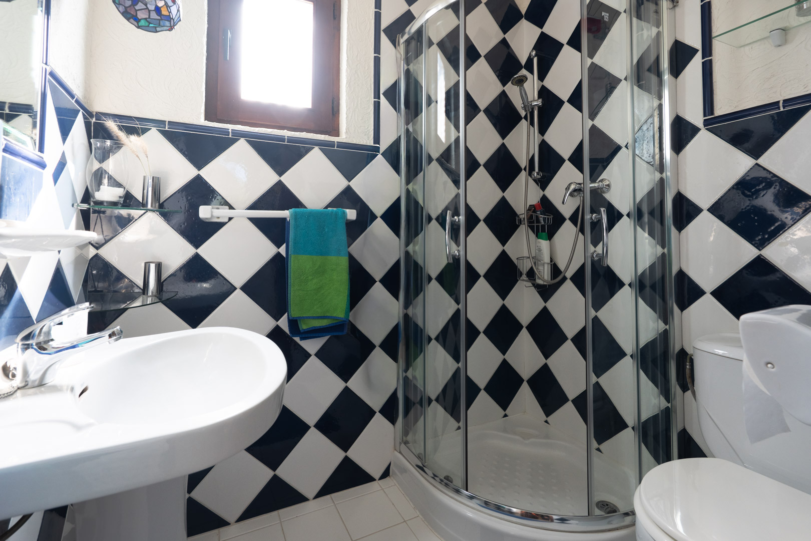 Badkamer met blauw-witte ruitvormige wandtegels, voorzien van een halfronde glazen douchecabine, een witte wastafel en een houten raamkozijn.