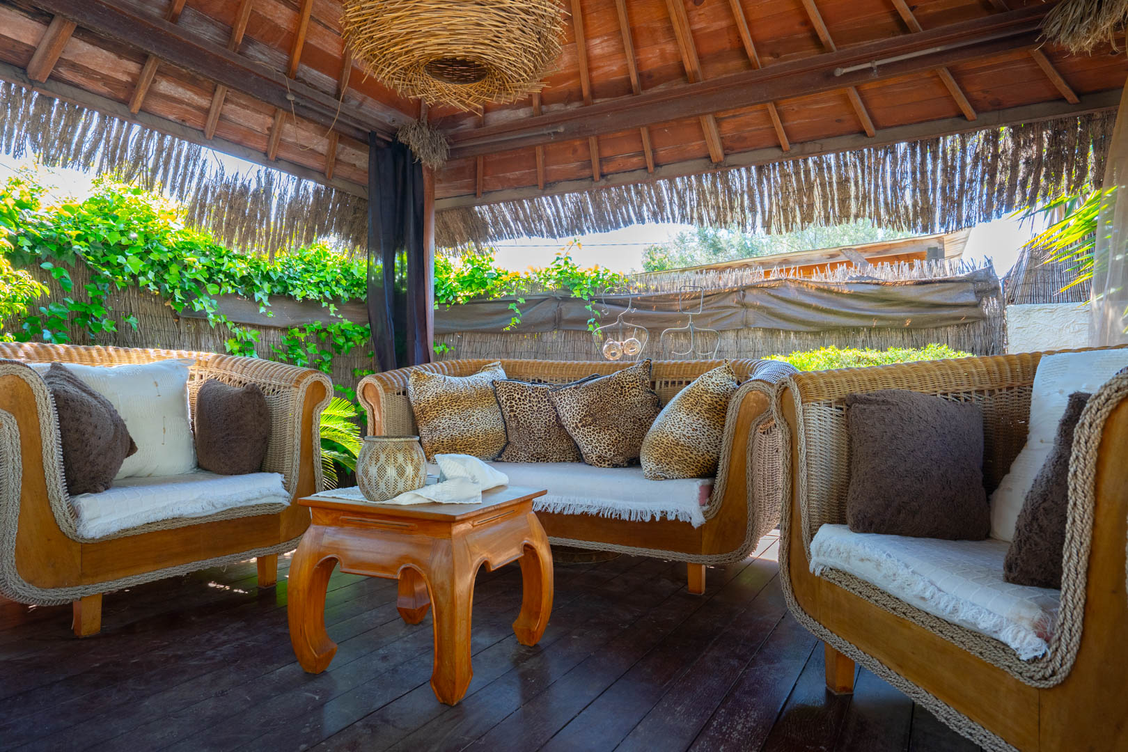 Balinese pergola met een houten balkenplafond, rieten loungemeubels met decoratieve kussens en een houten salontafel op een vlonderpad.