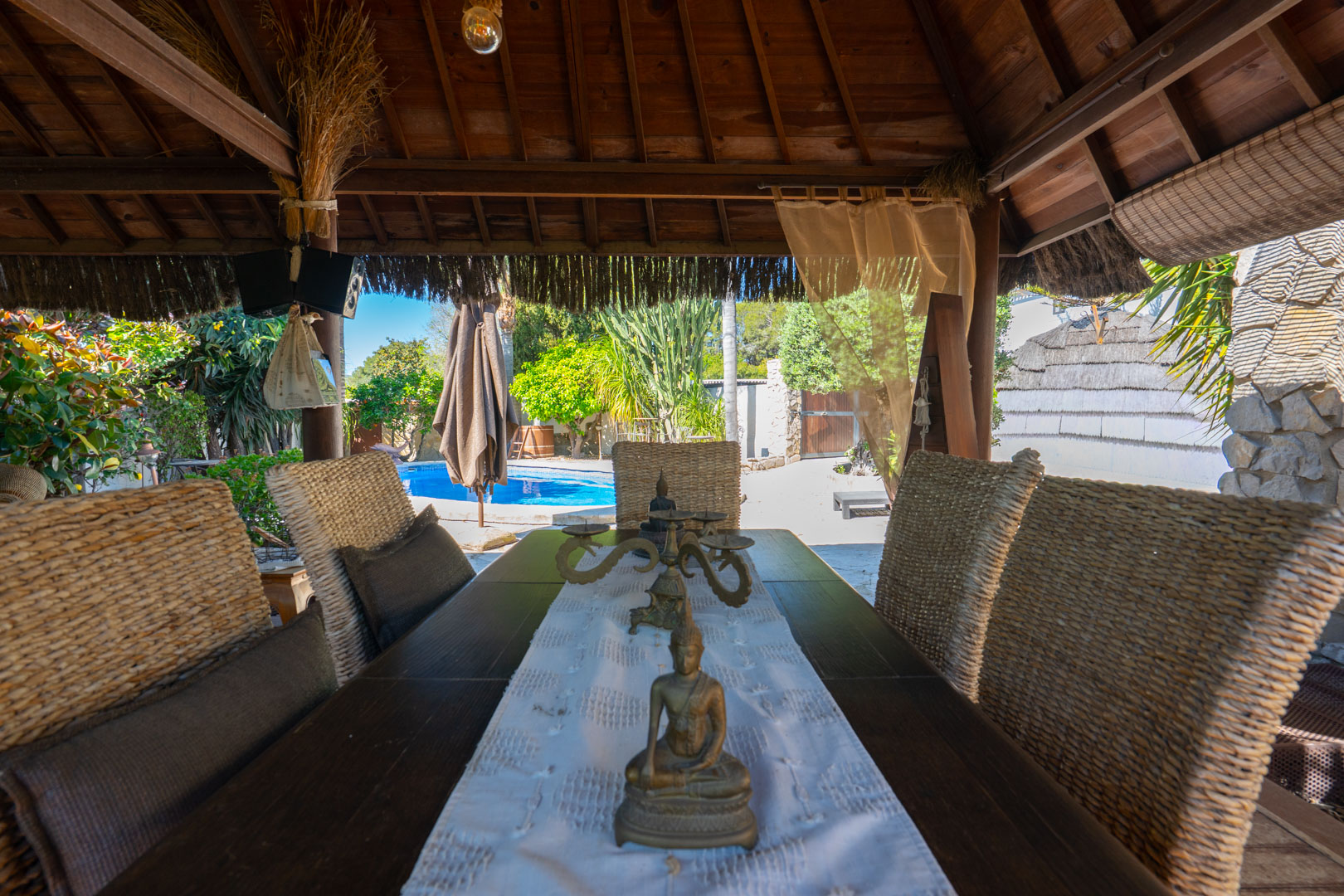 Buiten-eethoek onder een Balinese houten pergola, voorzien van rieten stoelen en uitzicht op het zwembad en de mediterrane tuin.