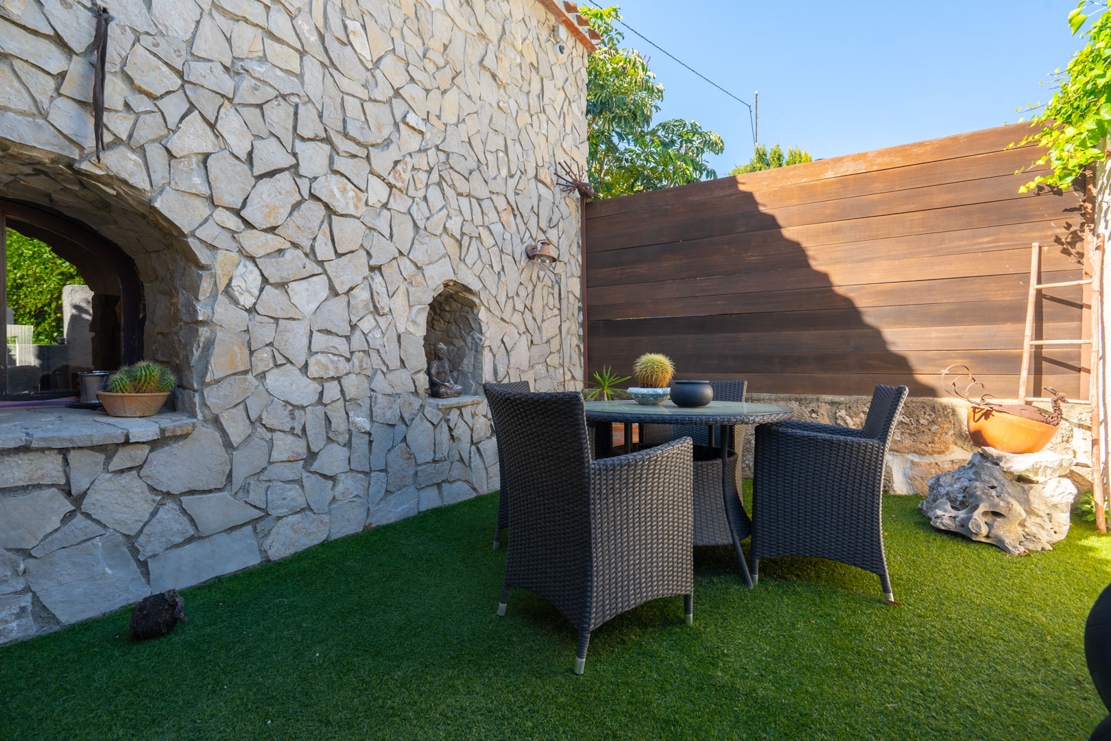 Buitenpatio met een natuurstenen muur voorzien van decoratieve nissen, een donkere rotan eethoek en een afwerking met kunstgras.