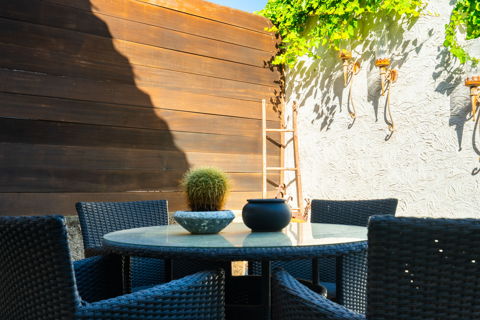 Buitenruimte met een ronde glazen tafel en donkere rotan stoelen, omgeven door een getextureerde muur met smeedijzeren kandelaars en houten schutting.