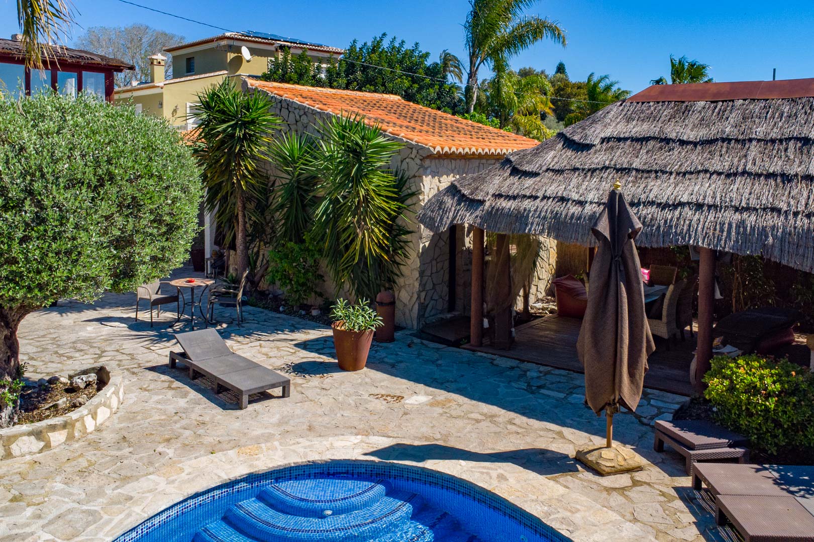 Buitenruimte met natuurstenen terras, zwembad en een Balinese pergola met rieten dak, gelegen naast een rustiek stenen bijgebouw en mediterrane beplanting.