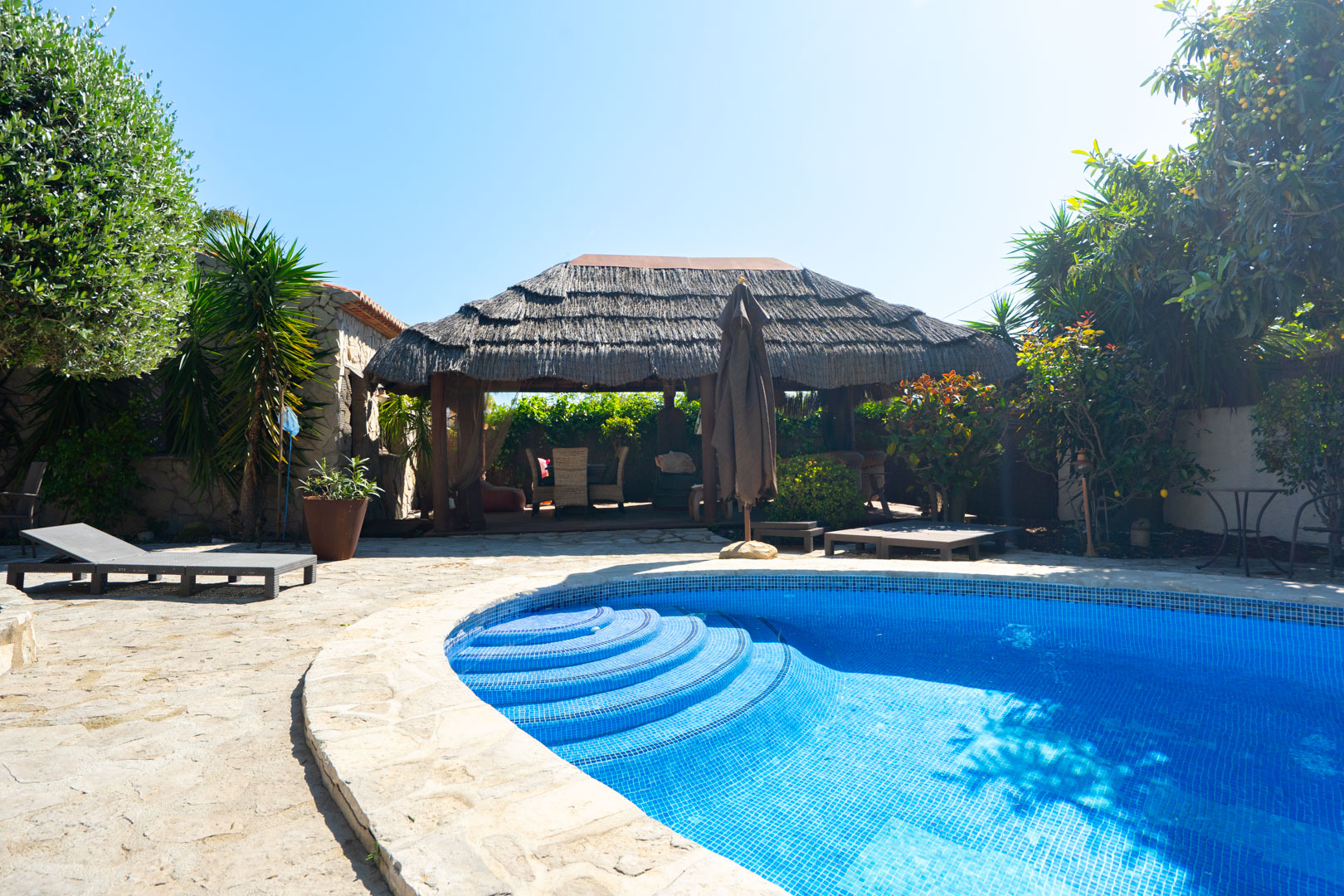 Buitenzwembad met blauwe mozaïektegels en ronde inlooptrap, omgeven door een natuurstenen terras en een grote Balinese pergola met rieten dak.