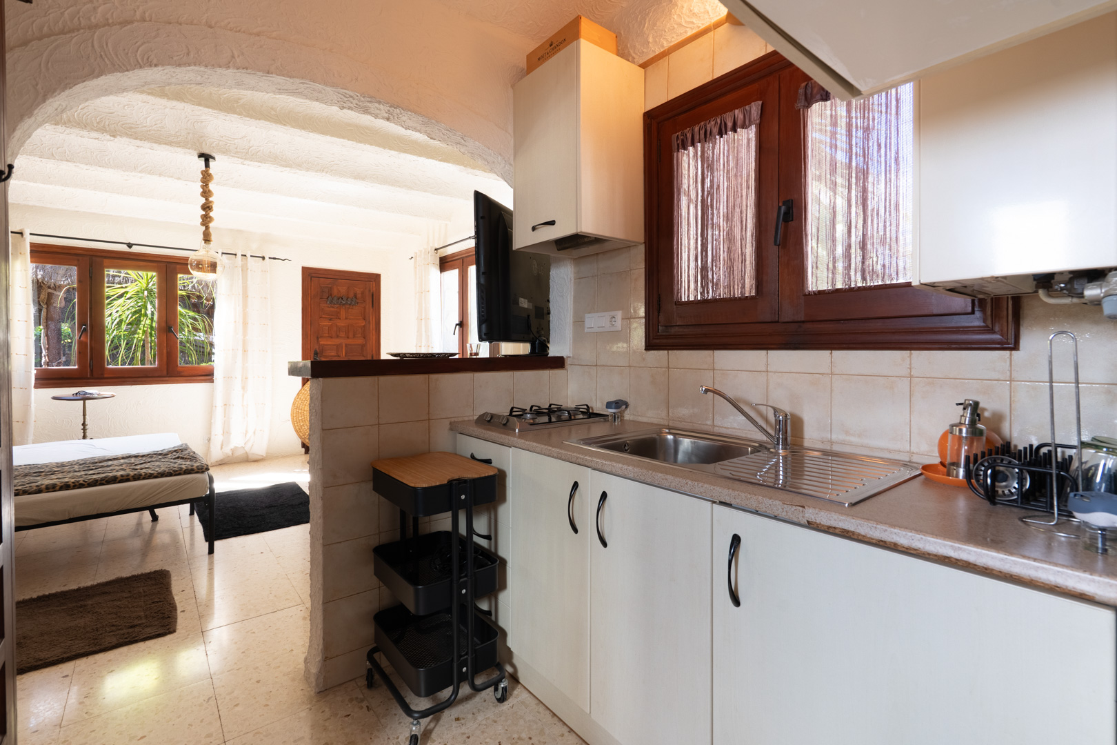 Compacte rustieke keuken met witte kasten en houten kozijnen, via een karakteristieke boog verbonden met een lichte slaapkamer.