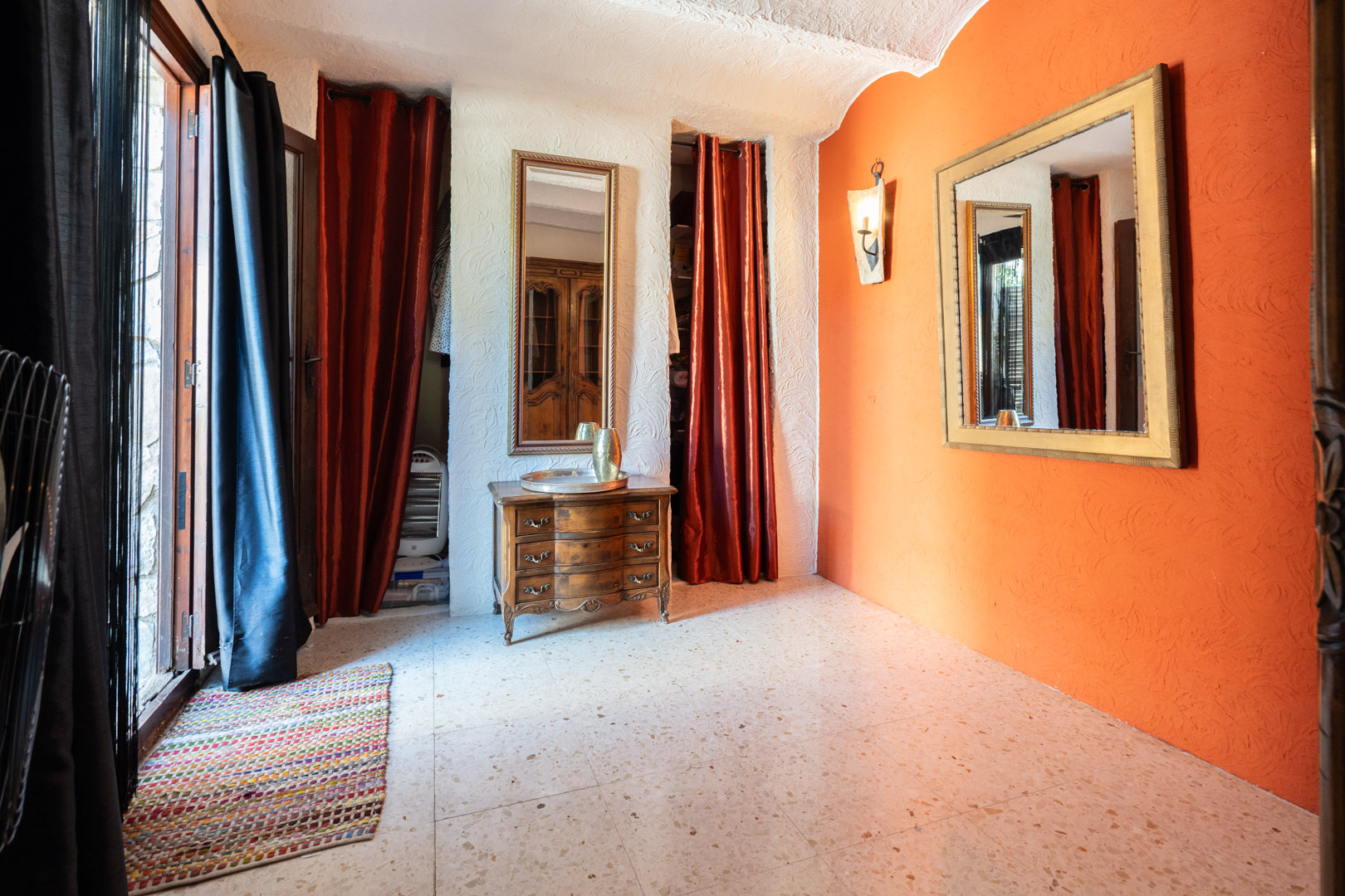Kleedruimte met een oranje accentmuur, sierlijke spiegels, een klassieke houten ladekast en terrazzovloer. Rode gordijnen schermen de ingebouwde kastruimtes af.