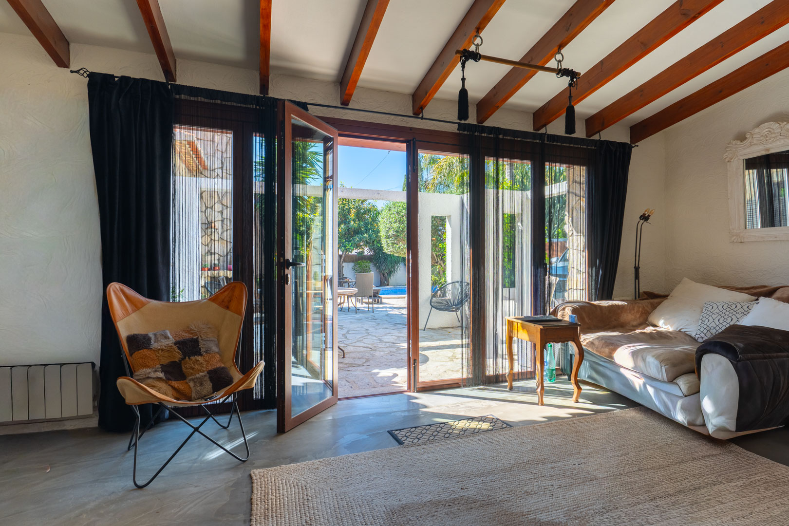 Lichte woonkamer met houten plafondbalken en grote glazen deuren die direct toegang bieden tot het stenen terras en het zwembad.