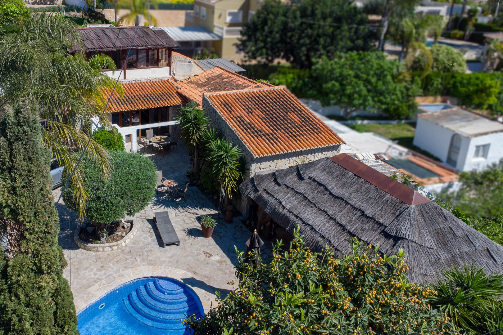 Luchtfoto van de rustieke villa met terracotta daken, een rond blauw betegeld zwembad, ruime natuurstenen terrassen en een grote Balinese pergola met rieten dak.