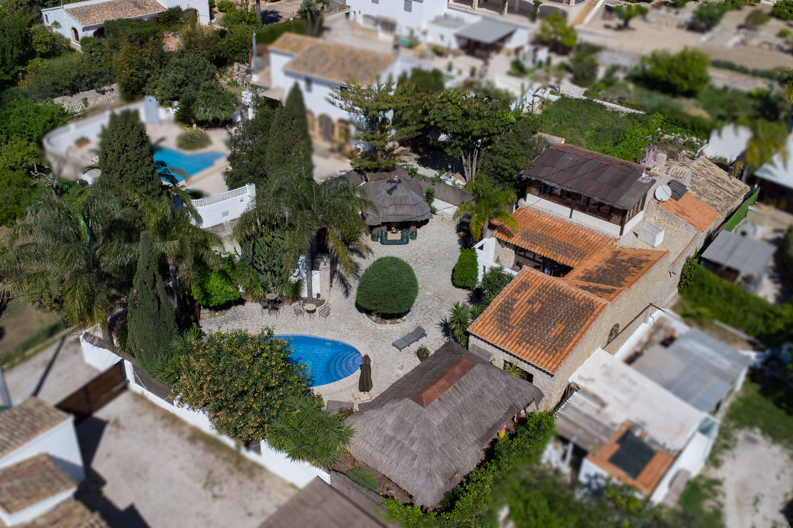 Luchtfoto van de villa met het ovale zwembad, natuurstenen terras en Balinese pergola's, omringd door dichte mediterrane beplanting en stenen muren.