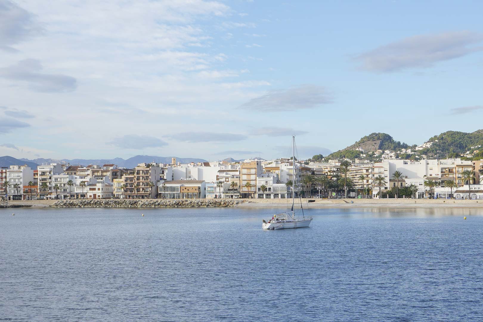 Panoramisch uitzicht op de haven van Jávea met een zeilboot voor de kust, omgeven door mediterrane architectuur en groene bergen.
