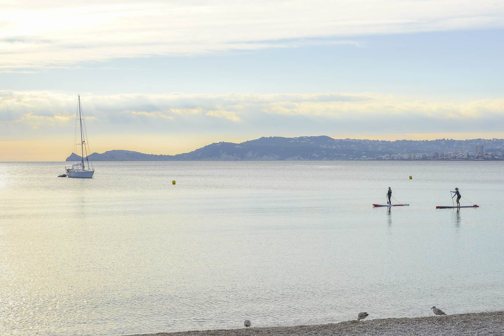 Panoramisch uitzicht over de rustige baai met een voor anker liggende zeilboot, suppers en de bergachtige kustlijn van Jávea.
