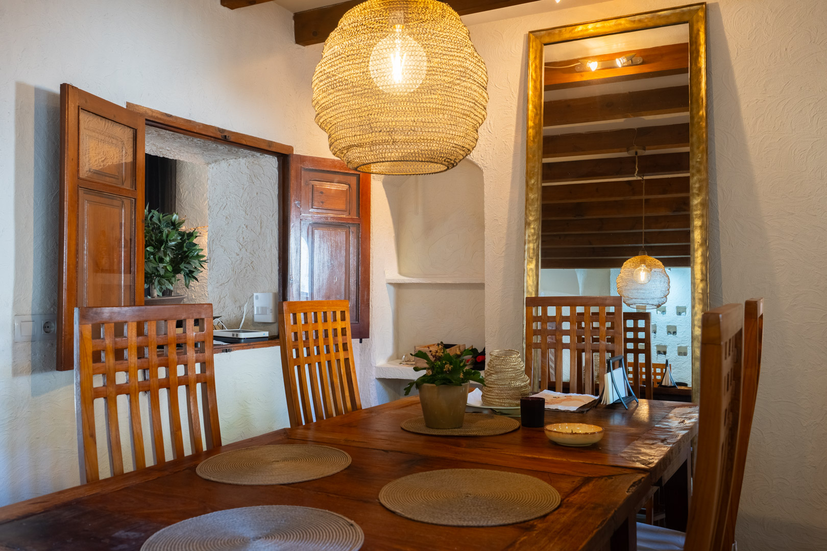 Rustieke eethoek met een massief houten tafel, stoelen met hoge rugleuning en een hanglamp van natuurlijke vezels. Voorzien van houten balken en een grote spiegel met gouden lijst.
