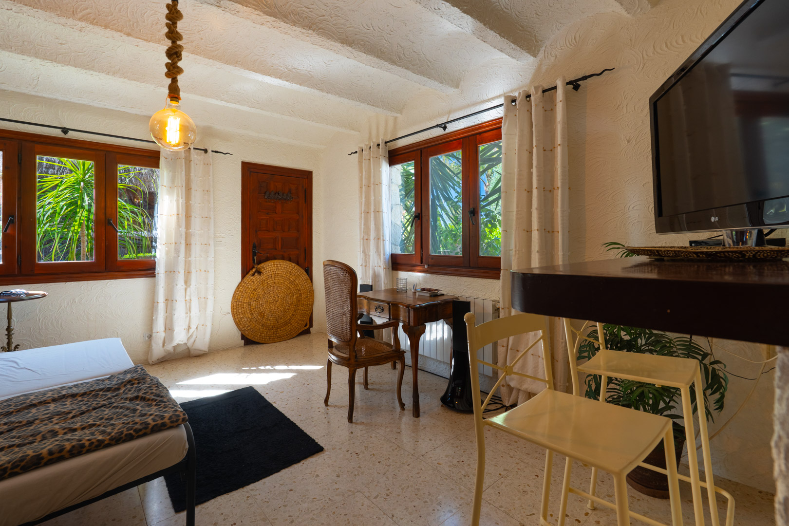 Rustieke logeerkamer met wit balkenplafond, klassiek houten bureau en ramen met donkere kozijnen die uitkijken op de mediterrane beplanting.