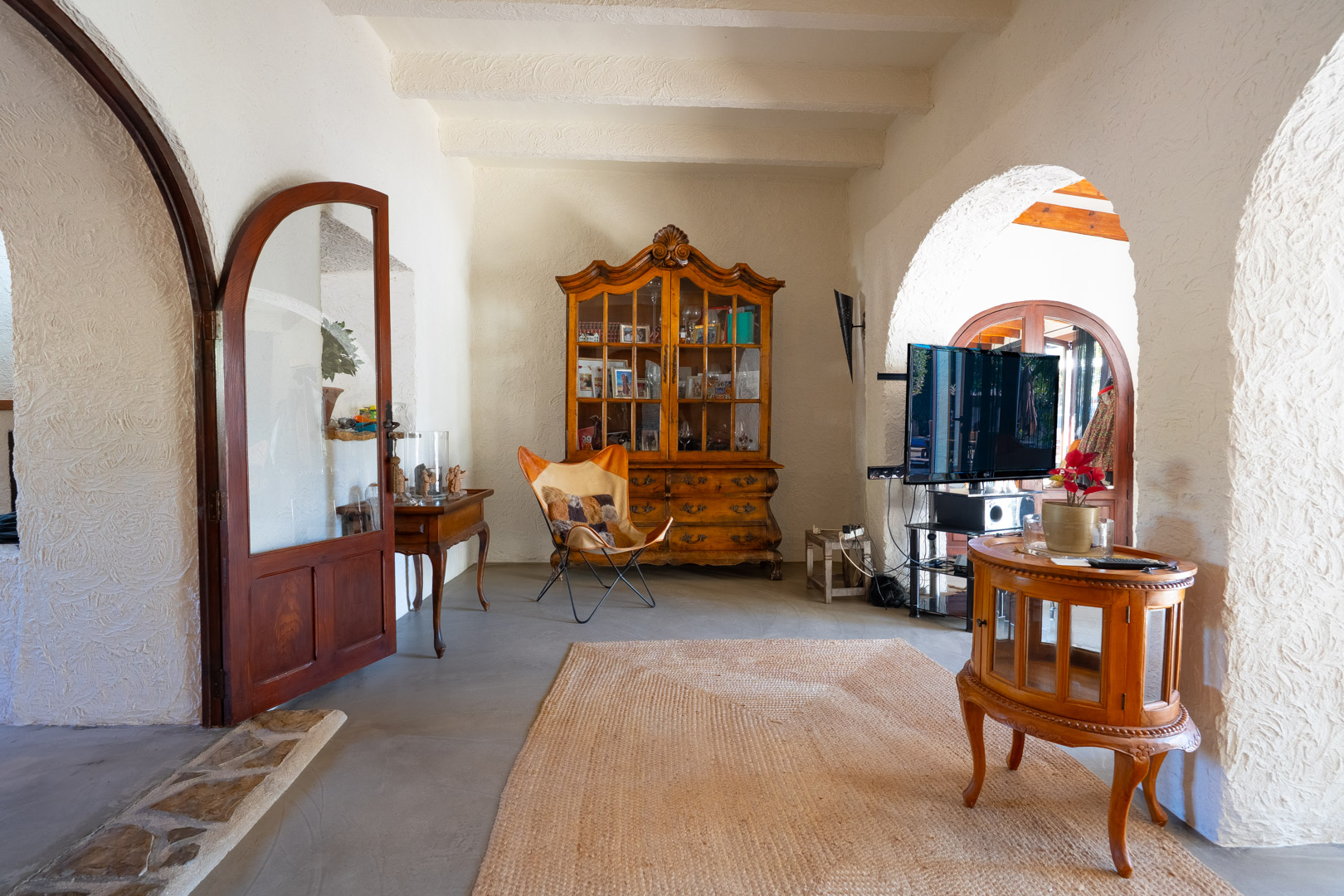 Woonkamer met witbalken plafonds, traditionele bogen en een microcement vloer. Voorzien van een antieke houten vitrinekast en rustiek meubilair.