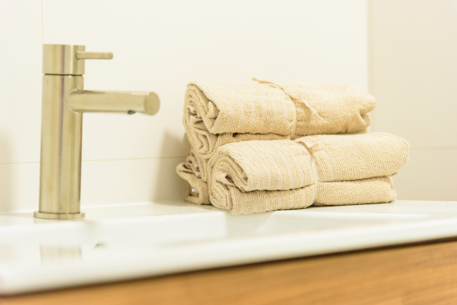 Detail van de badkamer met een geborsteld stalen kraan, witte wastafel en beige handdoeken op een modern houten badkamermeubel.