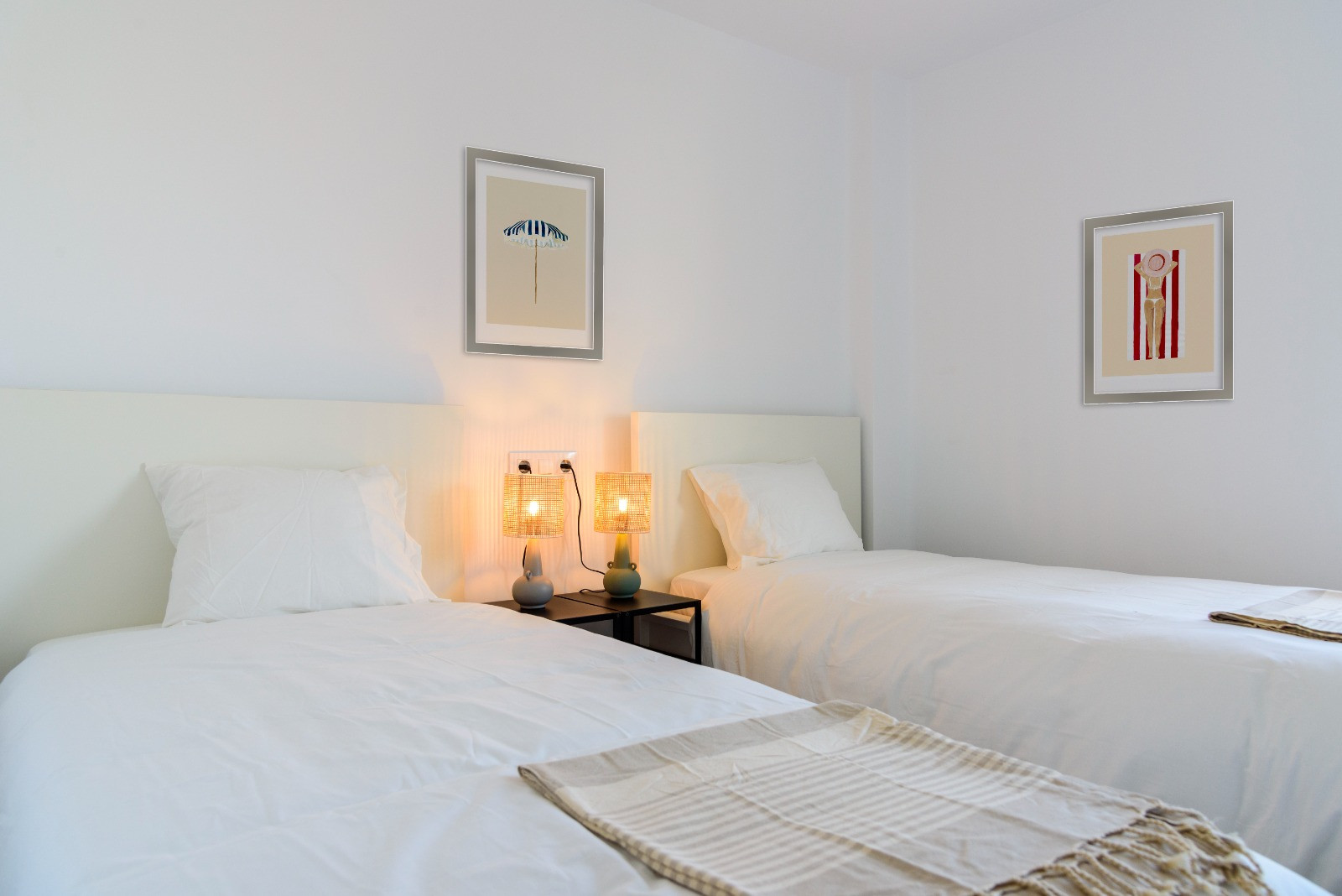 Lichte slaapkamer met twee eenpersoonsbedden, witte hoofdborden en lampen van natuurlijke vezels. Witte muren versierd met kunstwerken in een minimalistisch kustthema.