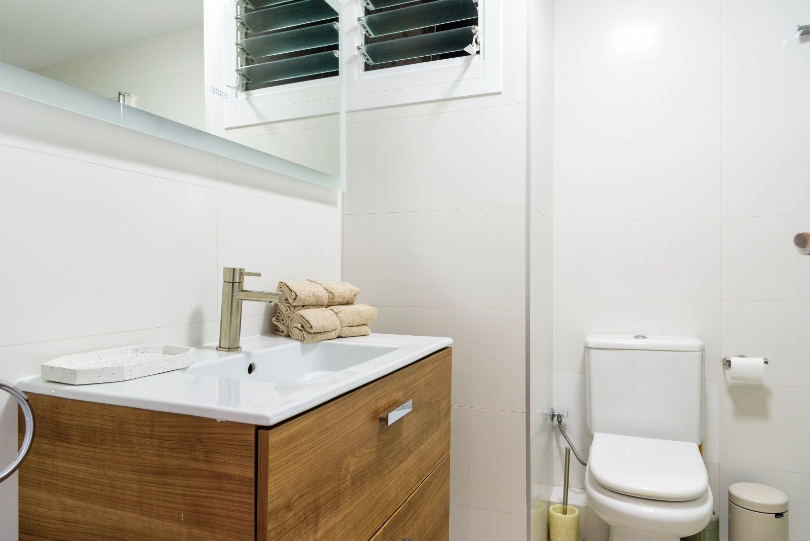 Moderne badkamer met houten wastafelmeubel, geïntegreerde wasbak en witte wandtegels. Voorzien van eigentijds sanitair en een lamellenvenster voor ventilatie.