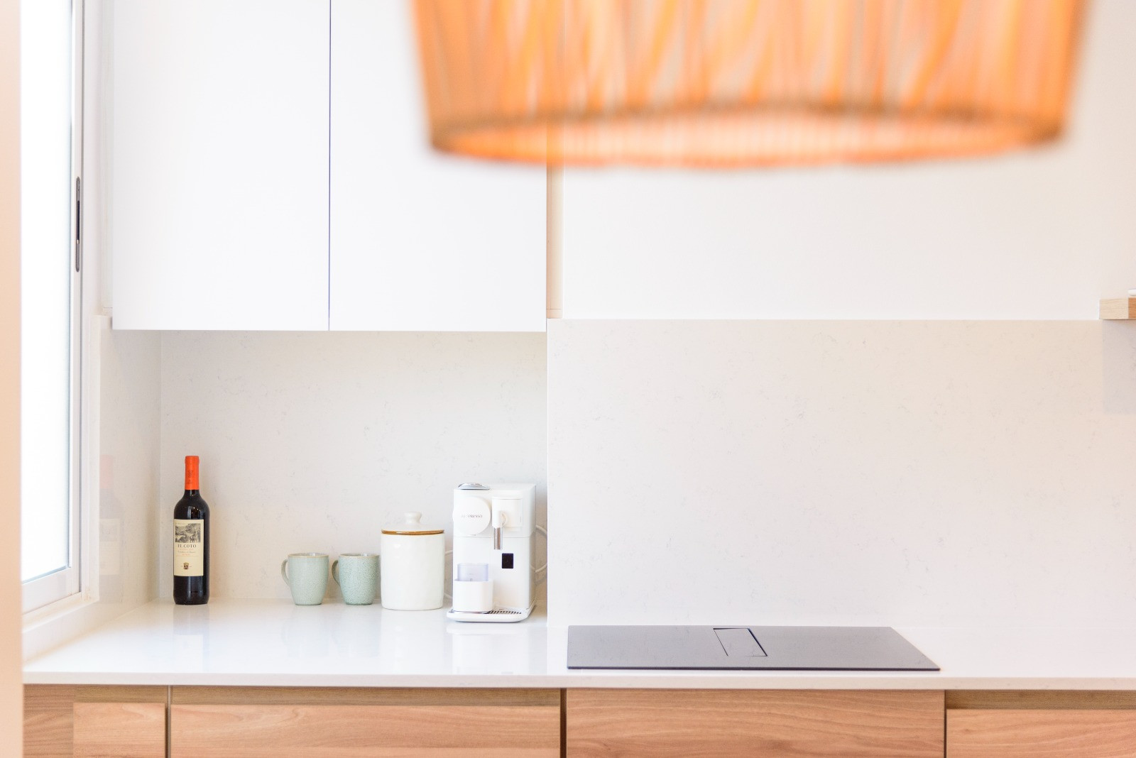 Moderne keuken met wit stenen werkblad, geïntegreerde inductiekookplaat en houten onderkasten. Natuurlijk licht verlicht de minimalistische koffiehoek en de strakke afwerking.