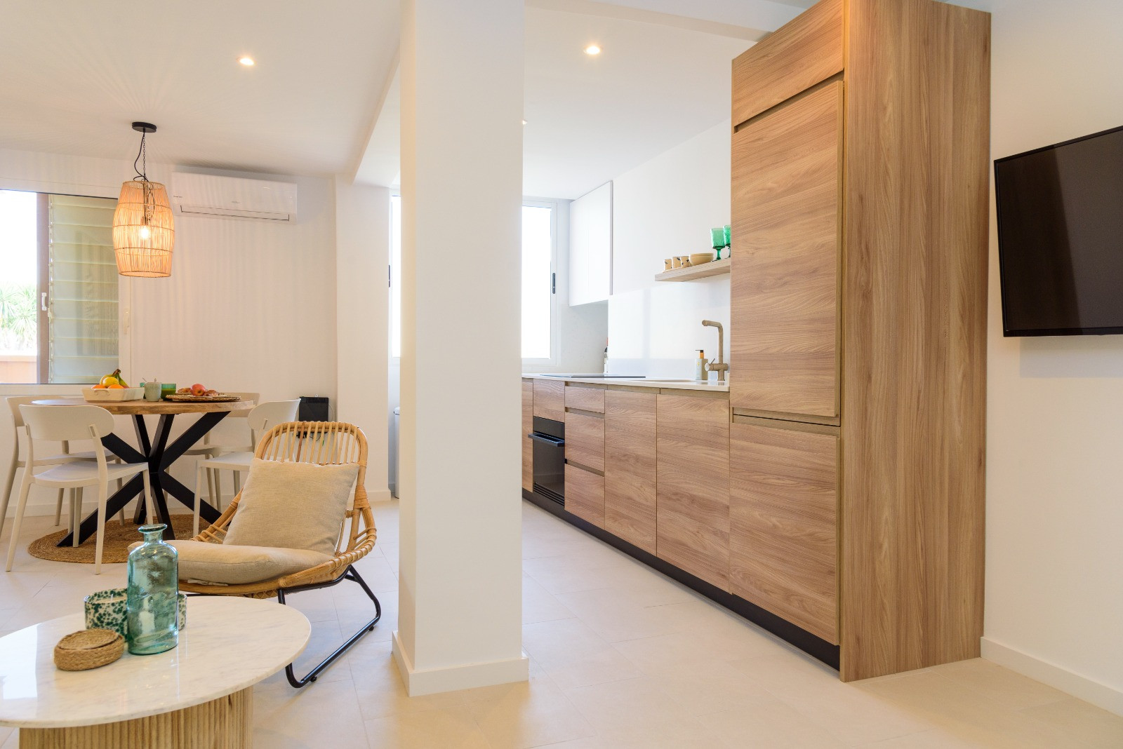 Open leefruimte met moderne houten keuken, eethoek met rotan hanglamp en lichte tegelvloer, afgewerkt met strakke witte muren.