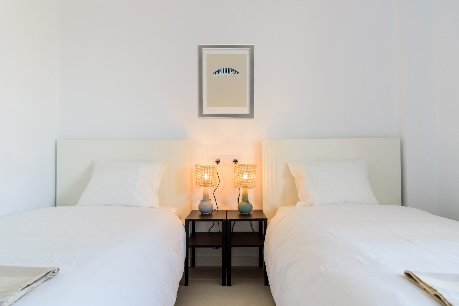 Slaapkamer met twee eenpersoonsbedden, witte hoofdborden en zwarte nachtkastjes voorzien van gevlochten lampen die een warme sfeer creëren in de lichte ruimte.