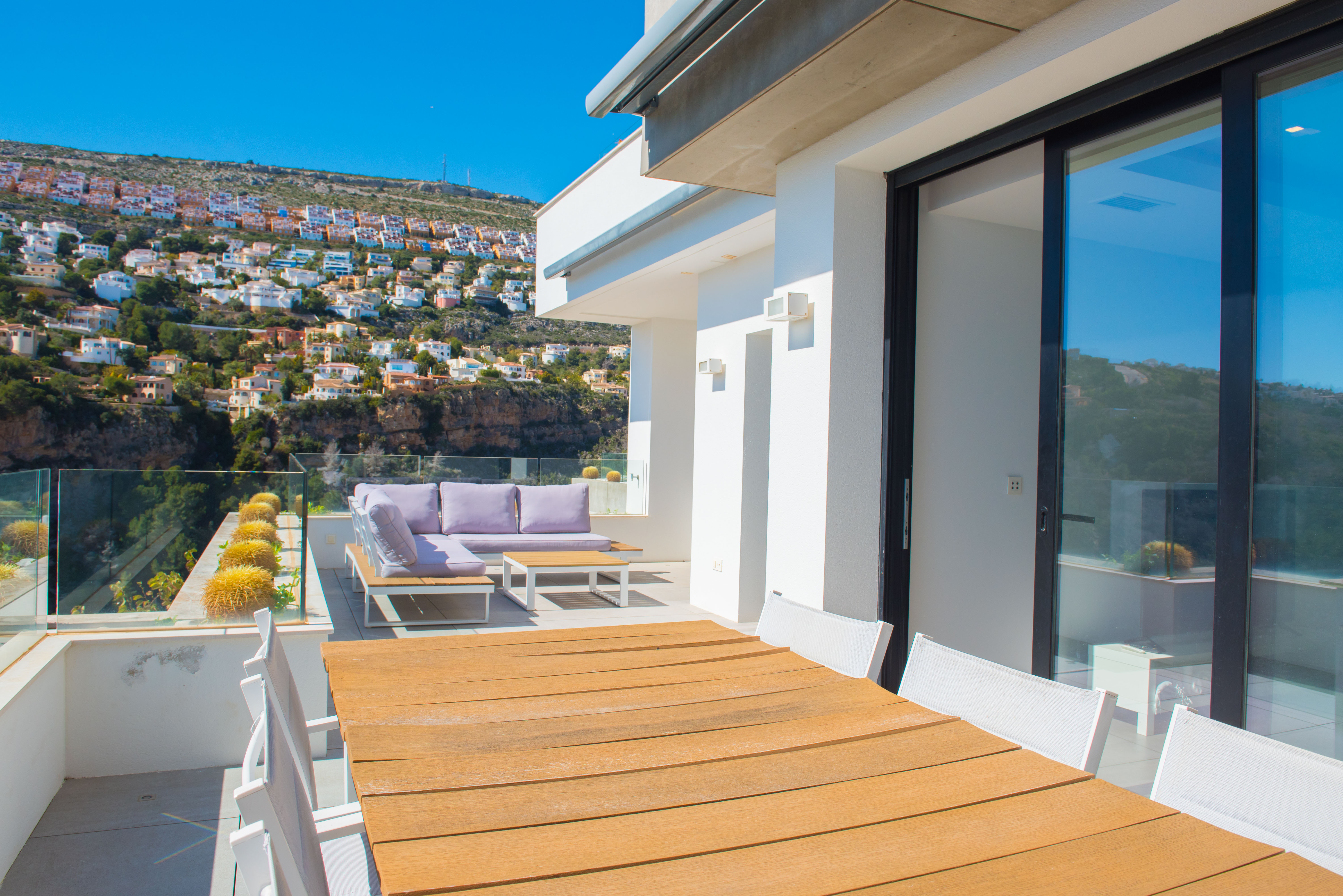 Buitenterras met houten eettafel, lila loungeset en glazen balustrades met uitzicht op de beboste heuvels en omliggende bebouwing.