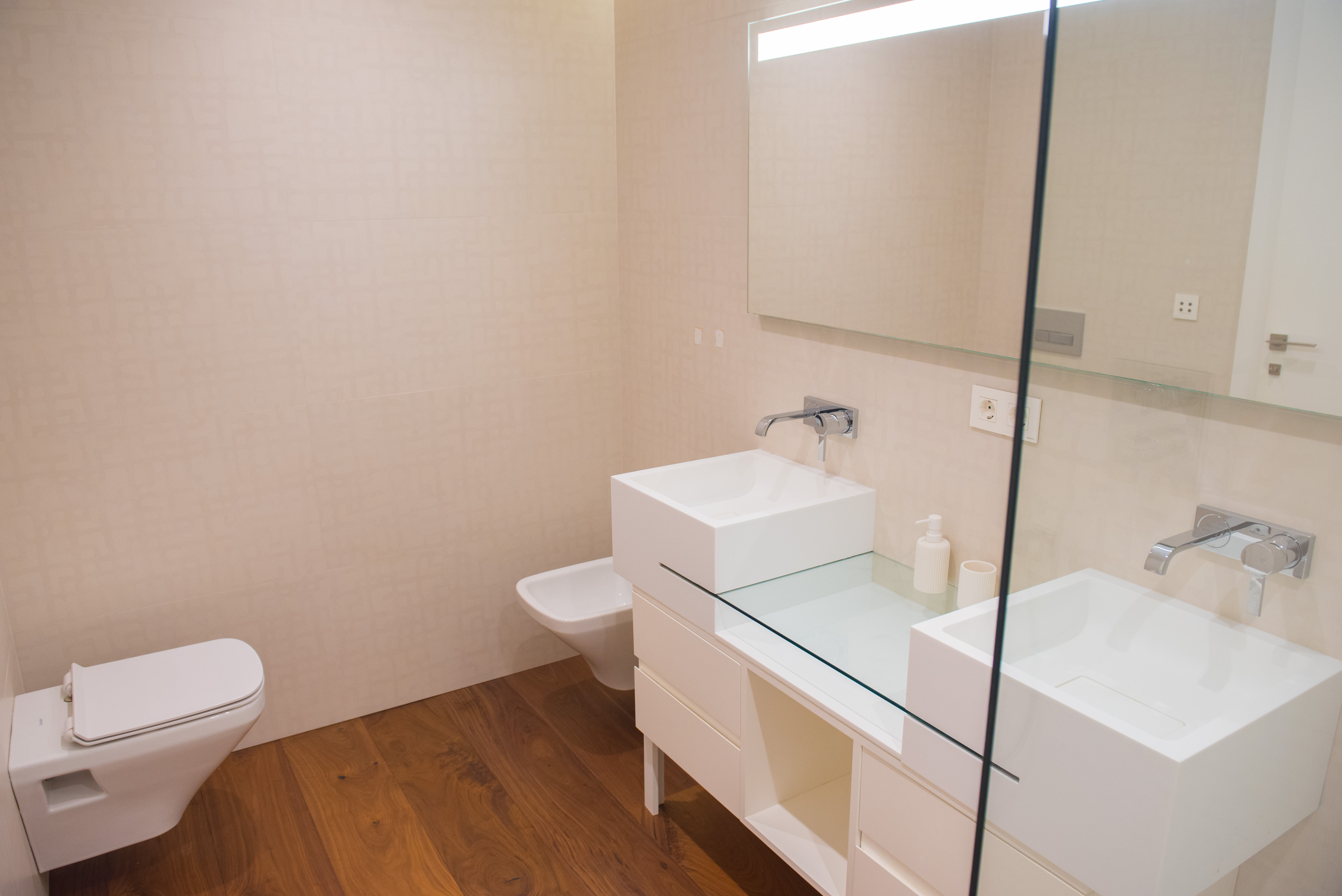 Eigentijdse badkamer met dubbele vierkante wastafels, glazen werkblad en wandkranen. Voorzien van hangend sanitair en een warme houten vloer.