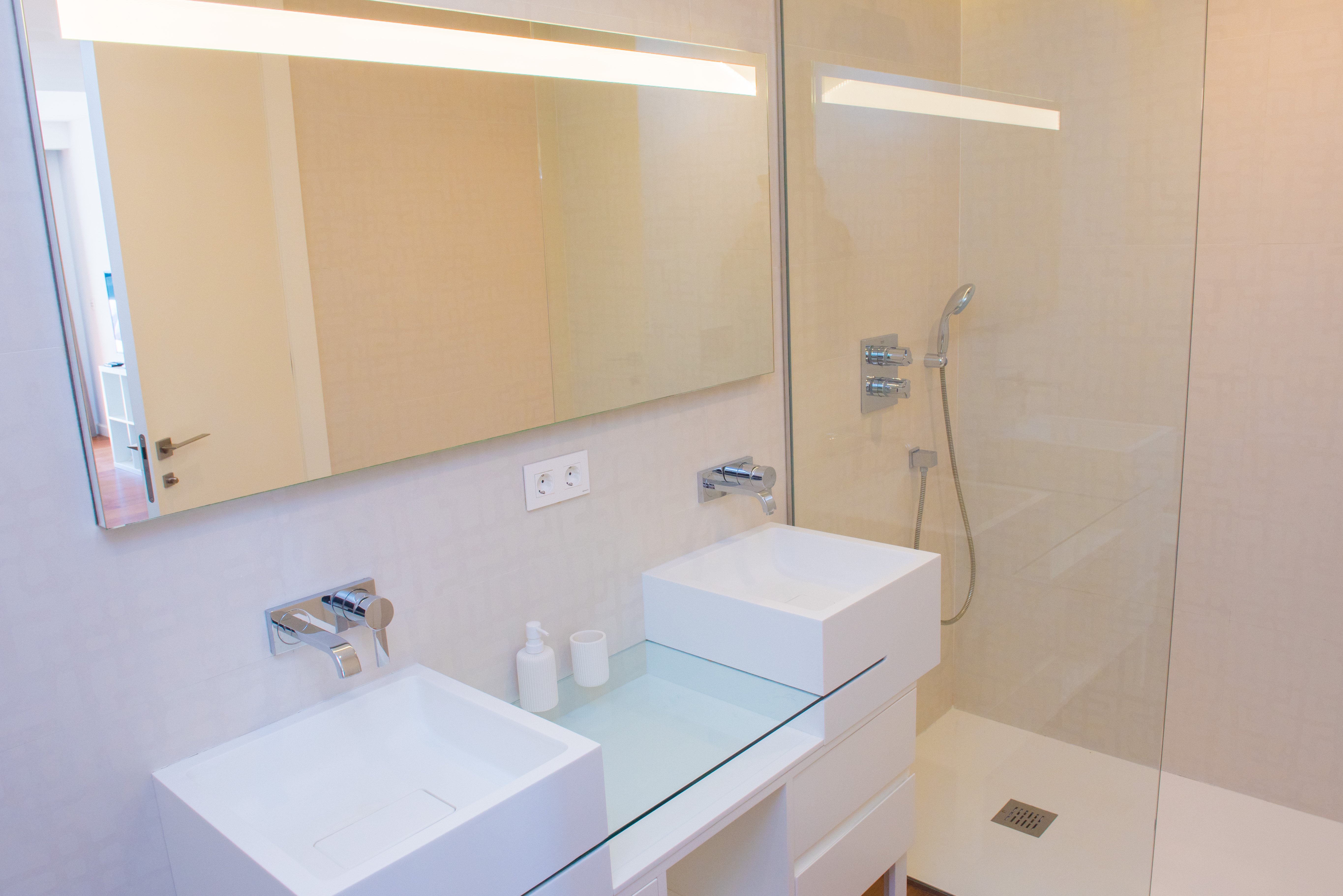 Eigentijdse badkamer met dubbele vierkante wastafels, inbouwkranen en een inloopdouche met glazen wand en spiegel met geïntegreerde LED-verlichting.