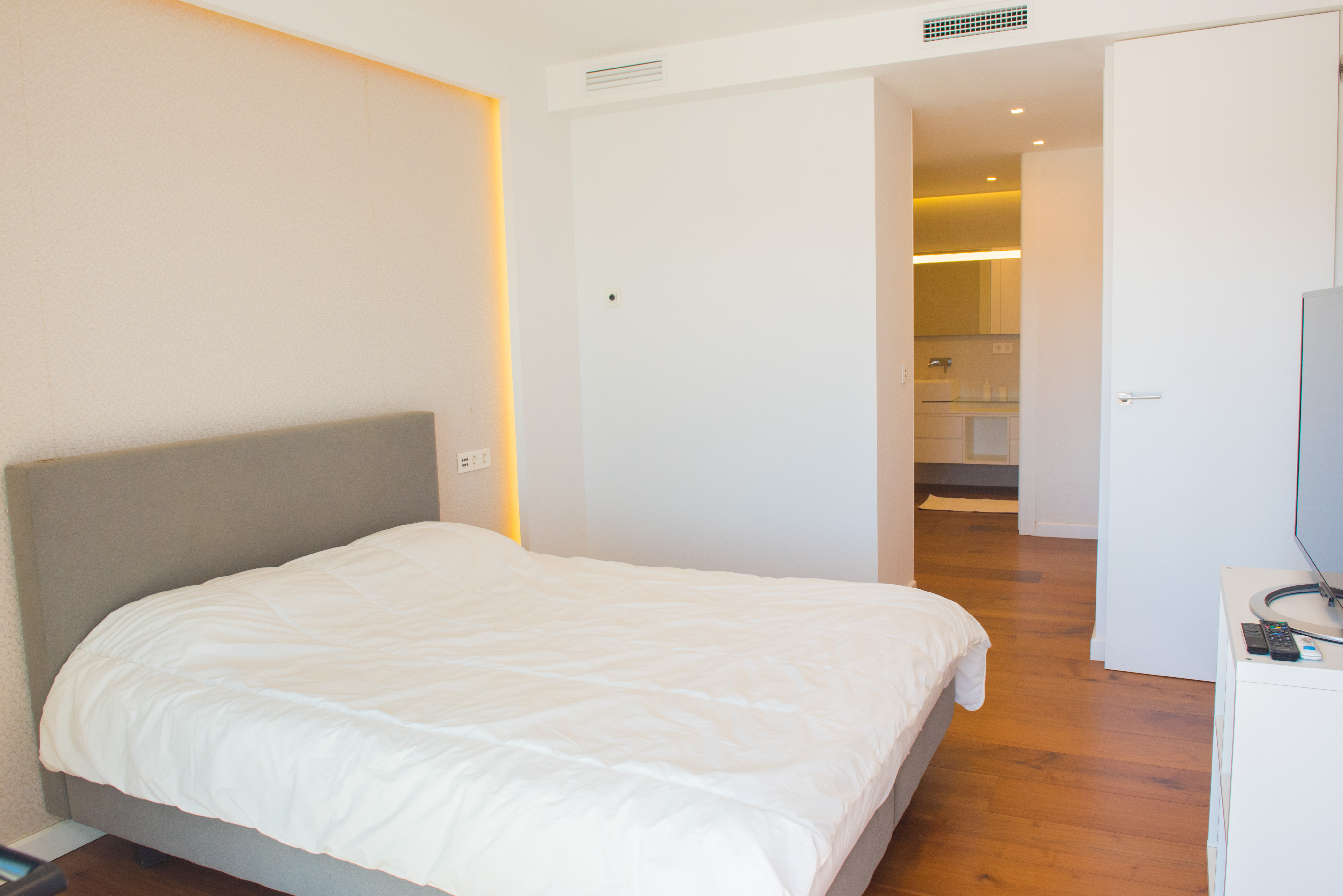Hoofdslaapkamer met grijs gestoffeerd hoofdbord, indirecte LED-wandverlichting, warme houten vloeren en doorkijk naar de moderne aangrenzende badkamer.