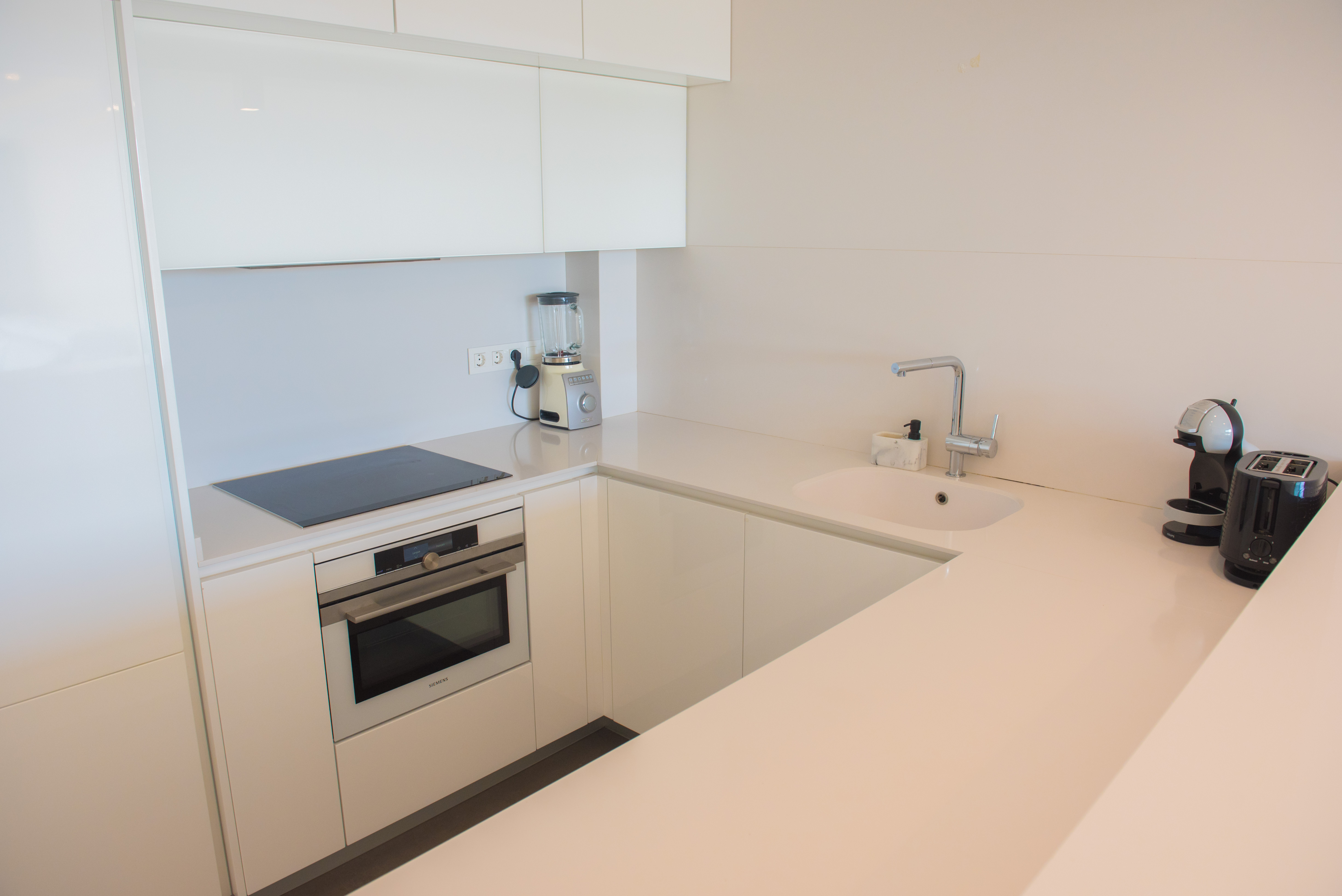 Minimalistische witglanzende keuken met kwarts werkblad, Siemens oven, inductiekookplaat en hoogwaardige keukenapparatuur in een praktische U-opstelling.