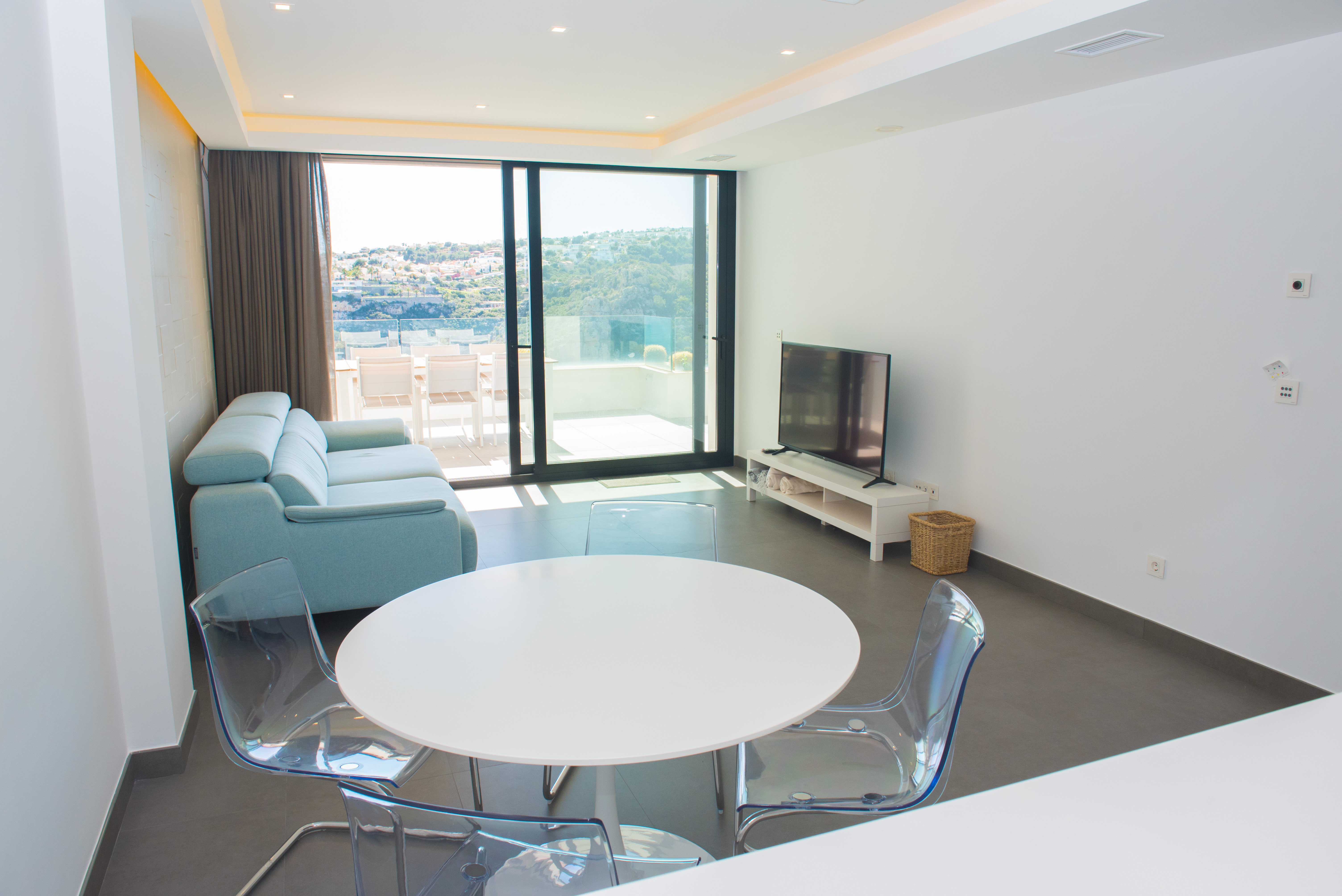 Minimalistische woon-eetkamer met witte ronde tafel, acryl stoelen en lichtblauwe bank, verbonden met een terras met panoramisch uitzicht via grote glazen schuifdeuren.