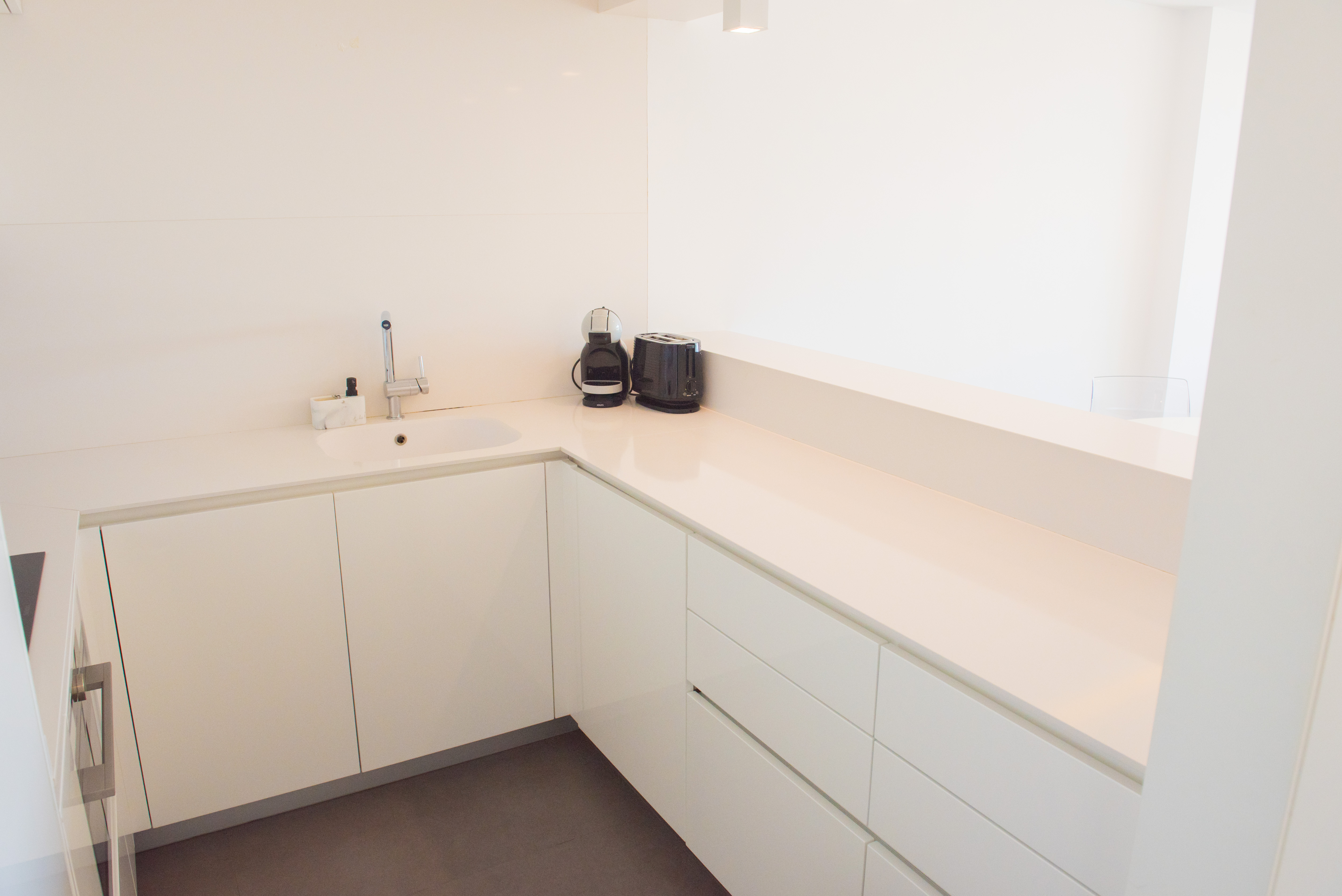 Moderne minimalistische keuken in hoogglans wit met een kwarts werkblad, geïntegreerde spoelbak en contrasterende donkere tegelvloer.